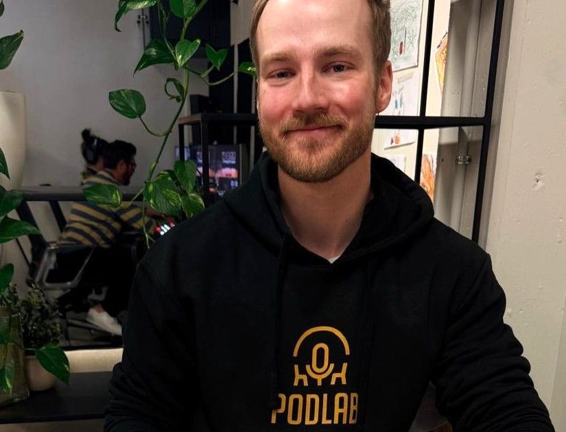 Smiling man in a black hoodie sitting indoors at a table, with plants and shelves in the background.