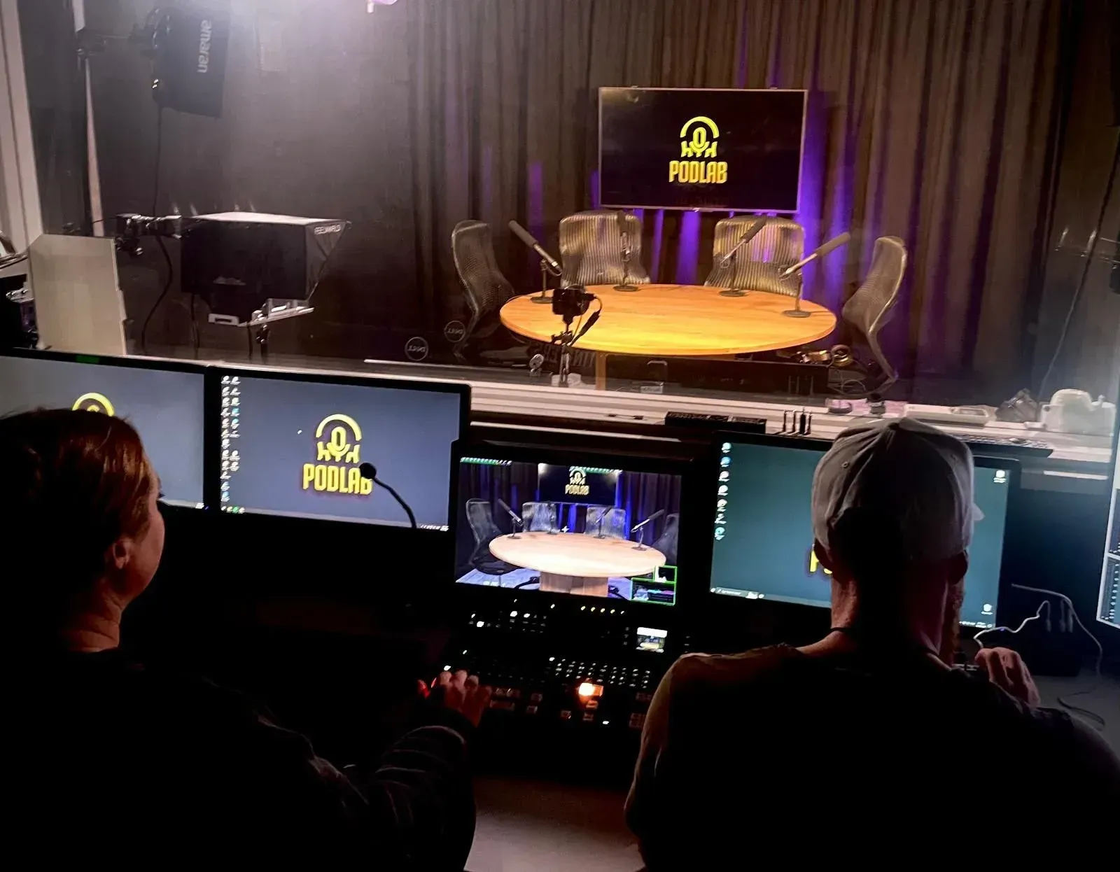 Control room view of a lit podcast set with three chairs and a monitor, seen from behind two operators