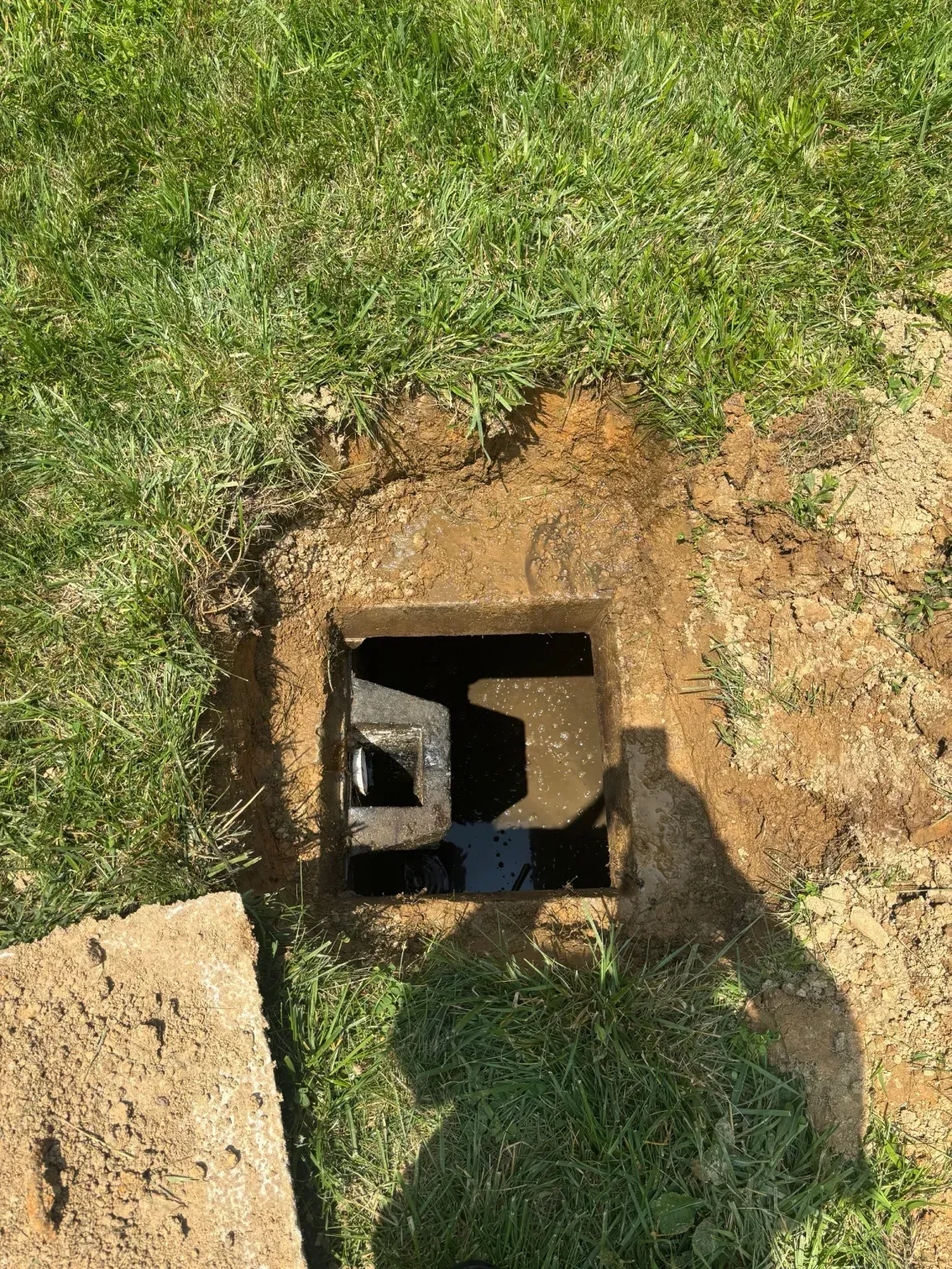 Open square utility hole in grass with a black pipe inside and a concrete slab nearby
