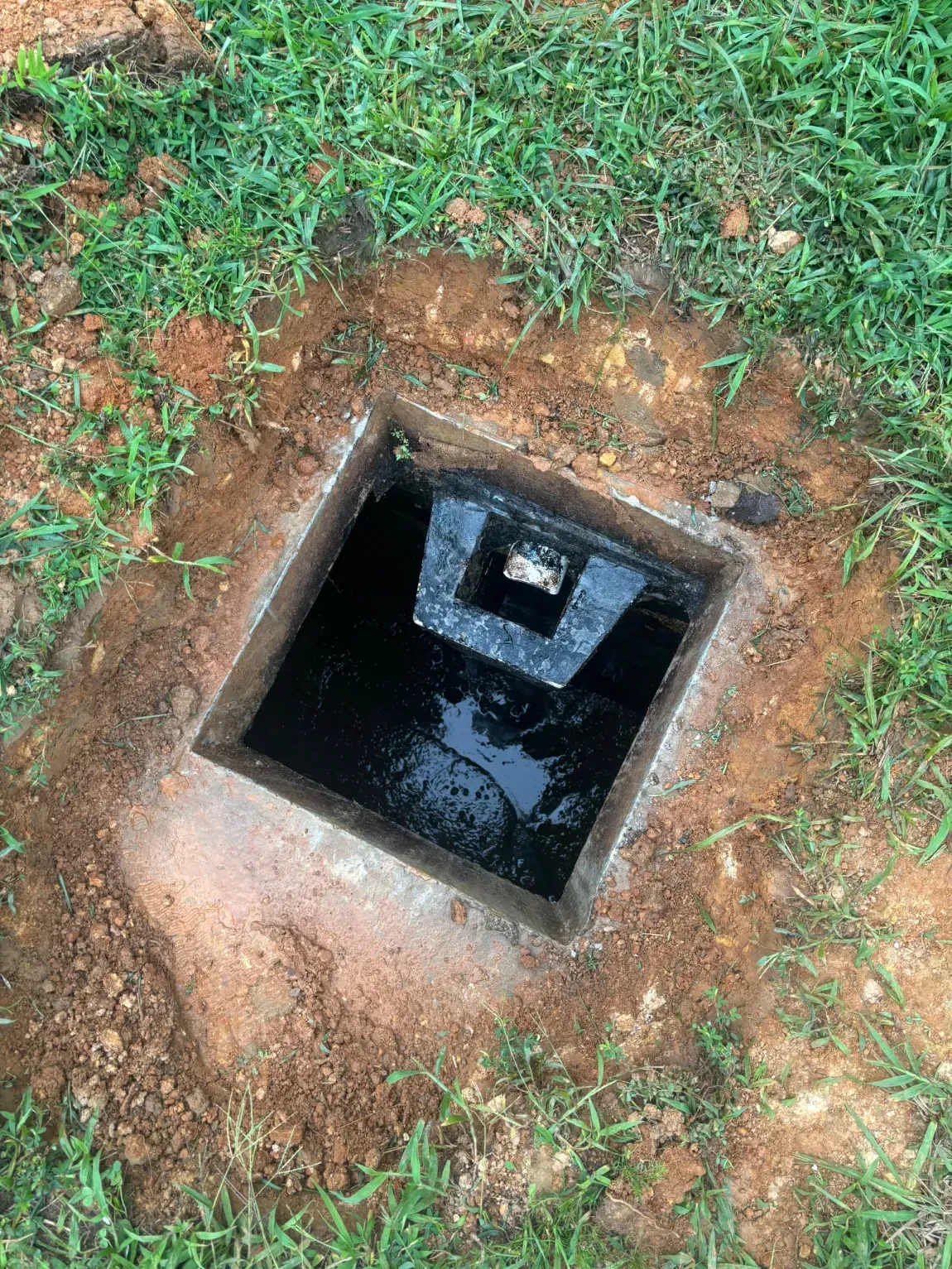 Square underground utility opening in grass, with a dark recessed shaft and metal frame.