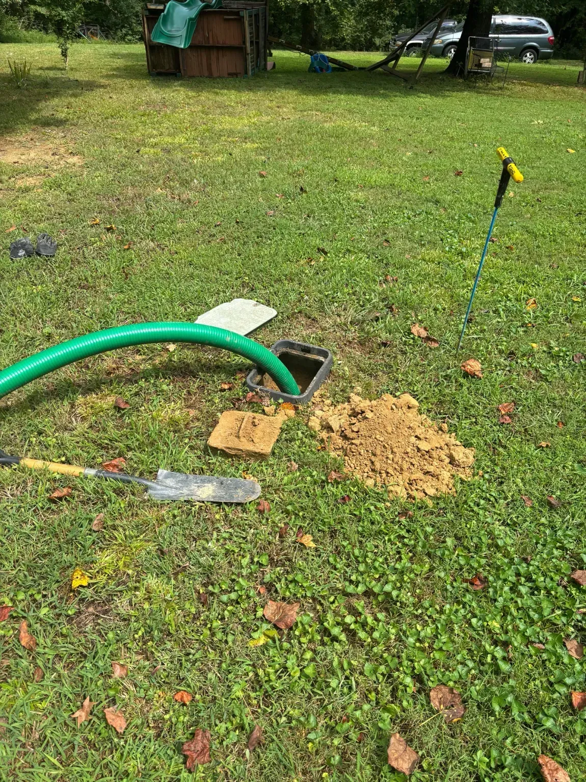 Lawn sprinkler setup in a grassy yard, with green hose, shovel, and a small dirt pile.
