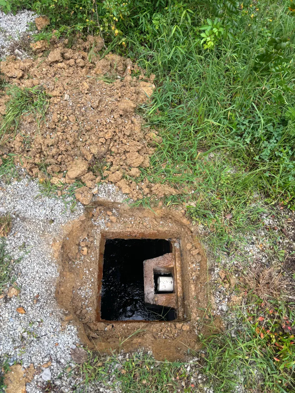 Open utility hole in grass and dirt beside a gravel path, with a pipe visible inside.
