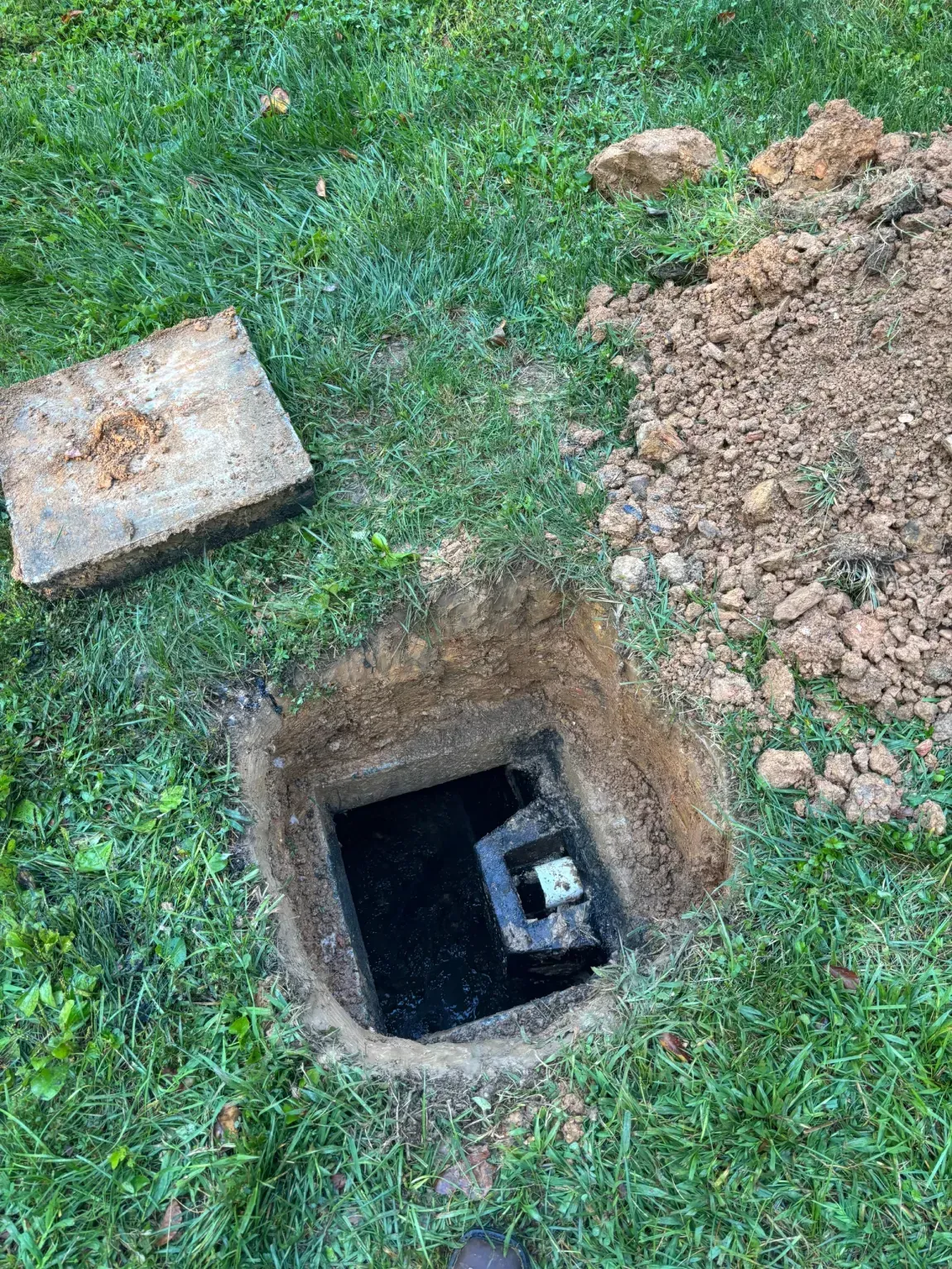 Open square utility hole in grassy yard beside a concrete cover and dirt pile