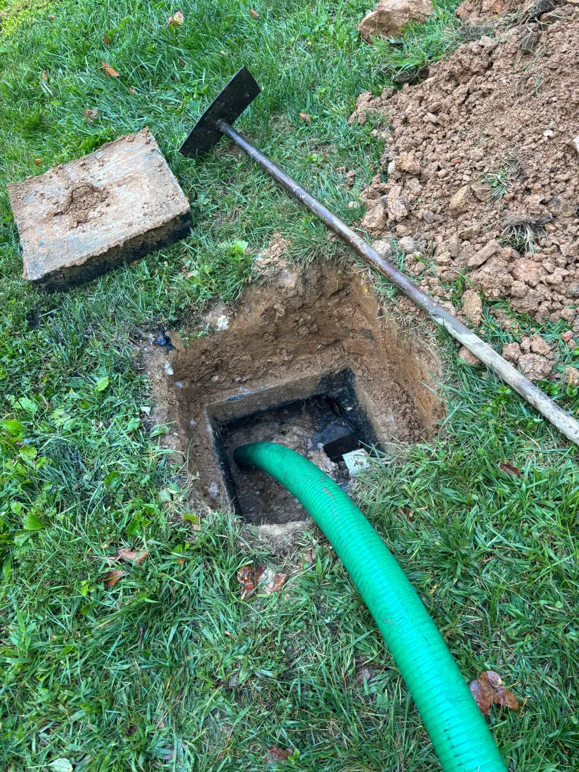 Green hose entering an underground utility box in a grassy yard, with a shovel nearby