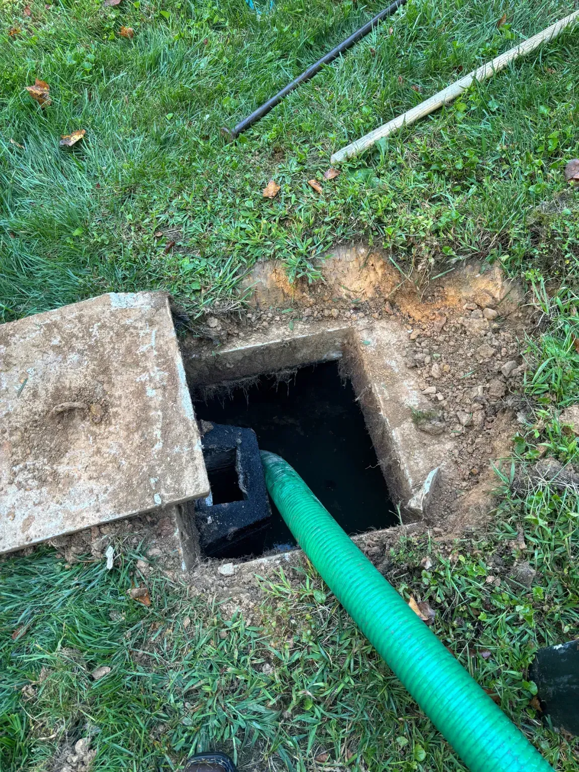 Open ground box with green hose entering a dark hole beside a concrete slab in grass