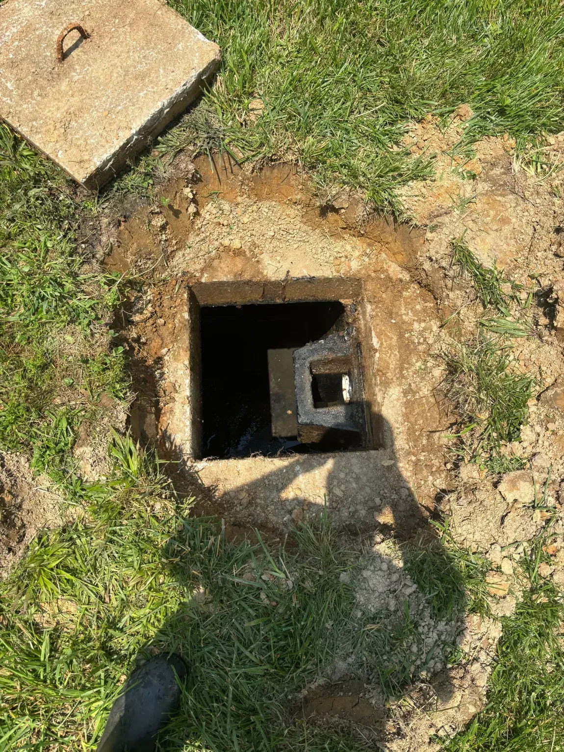 Open utility pit in grass with a metal fitting inside, viewed from above.