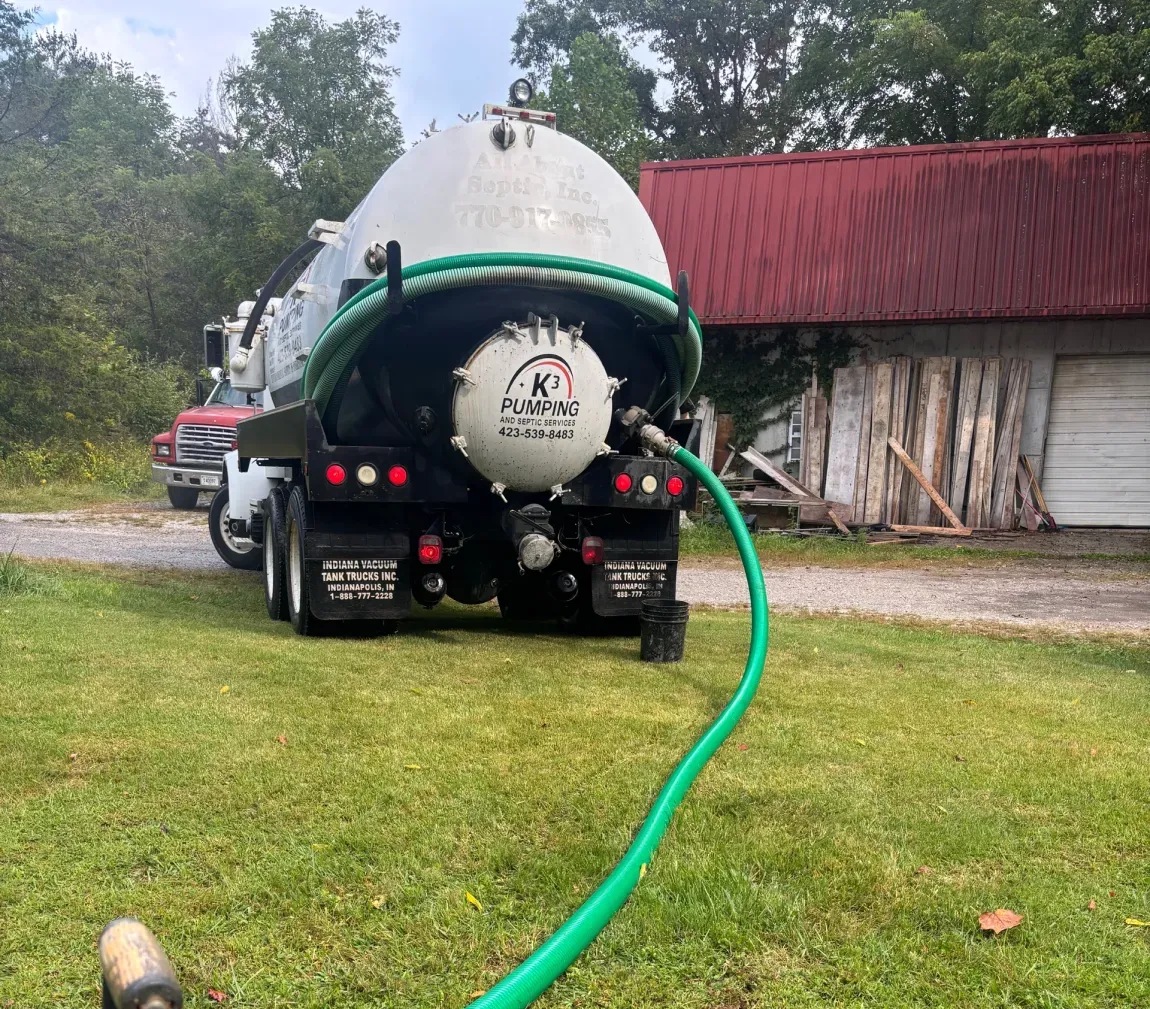 Septic tank truck on a grassy lawn, hose extended to a drain, beside a red-roofed building