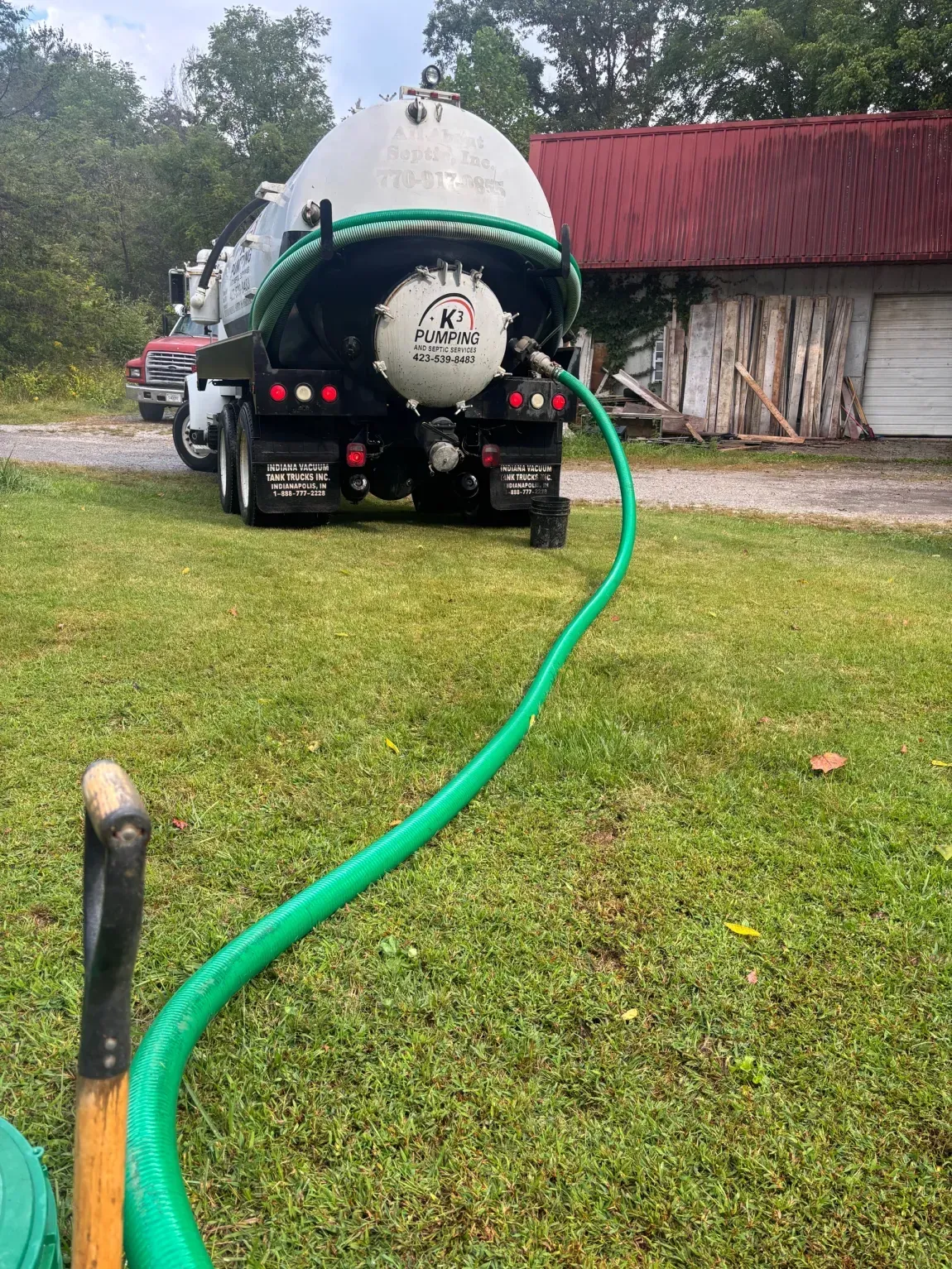 Green tanker truck on grass with a hose connected, parked beside a red-roofed barn.