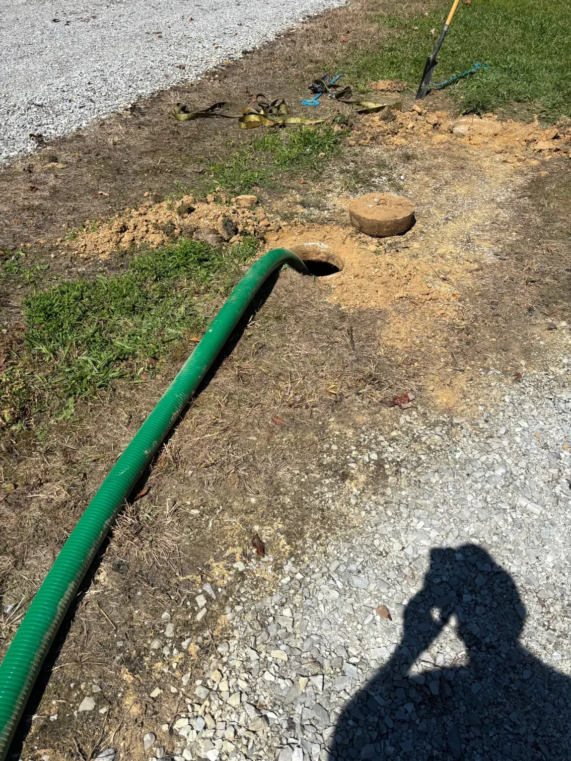 Green hose on gravel beside a dirt patch, with a round rock and a shadow in the foreground.