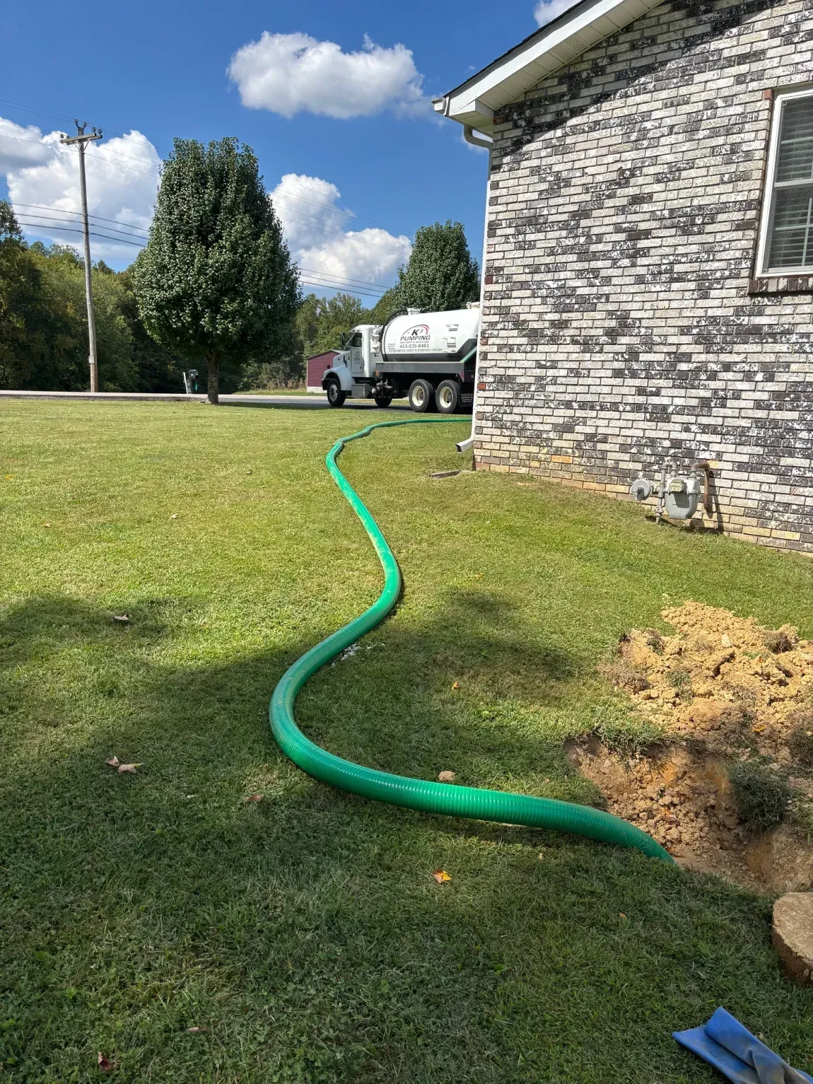Green hose on a lawn beside a stone house, leading toward a parked white truck under a blue sky.