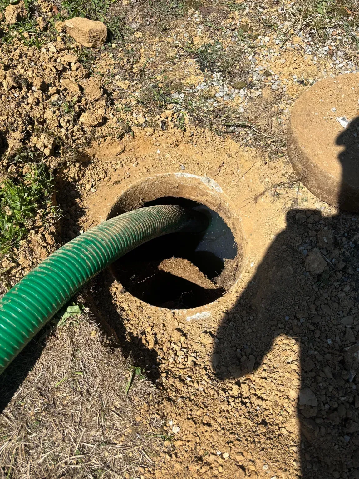 Green hose inserted into a circular ground opening surrounded by dirt and a shadow.