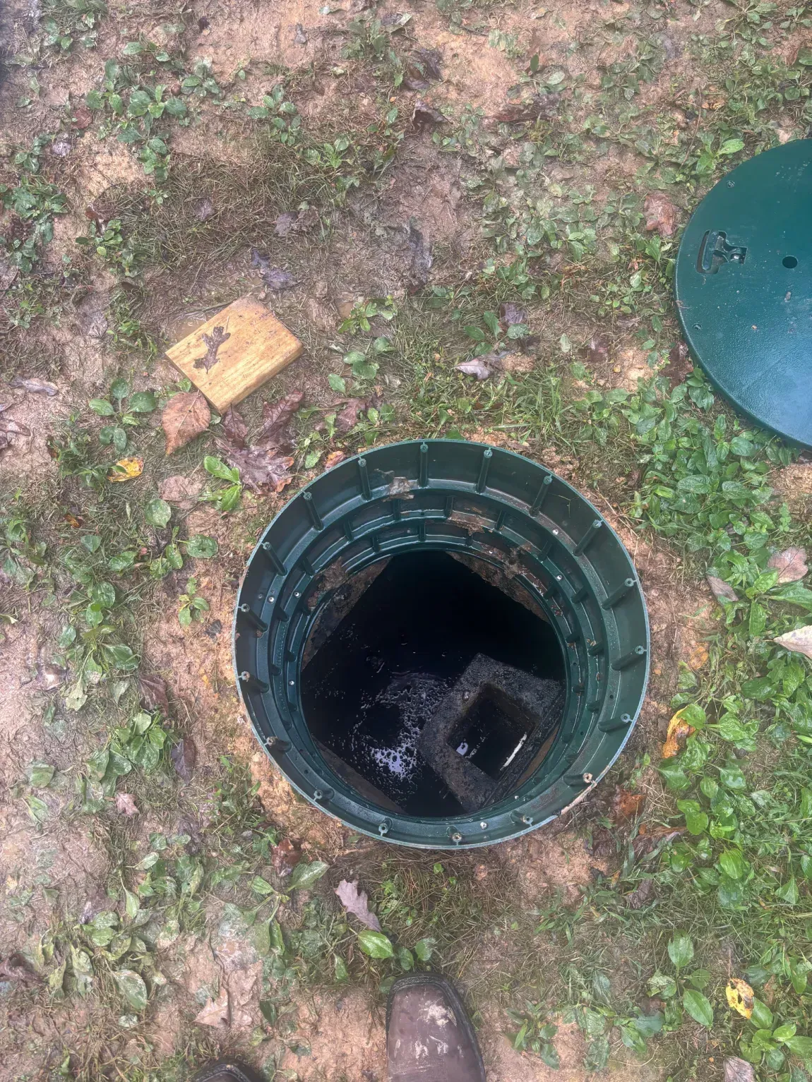 Open circular green access hole in grassy dirt ground with the cover partly visible nearby