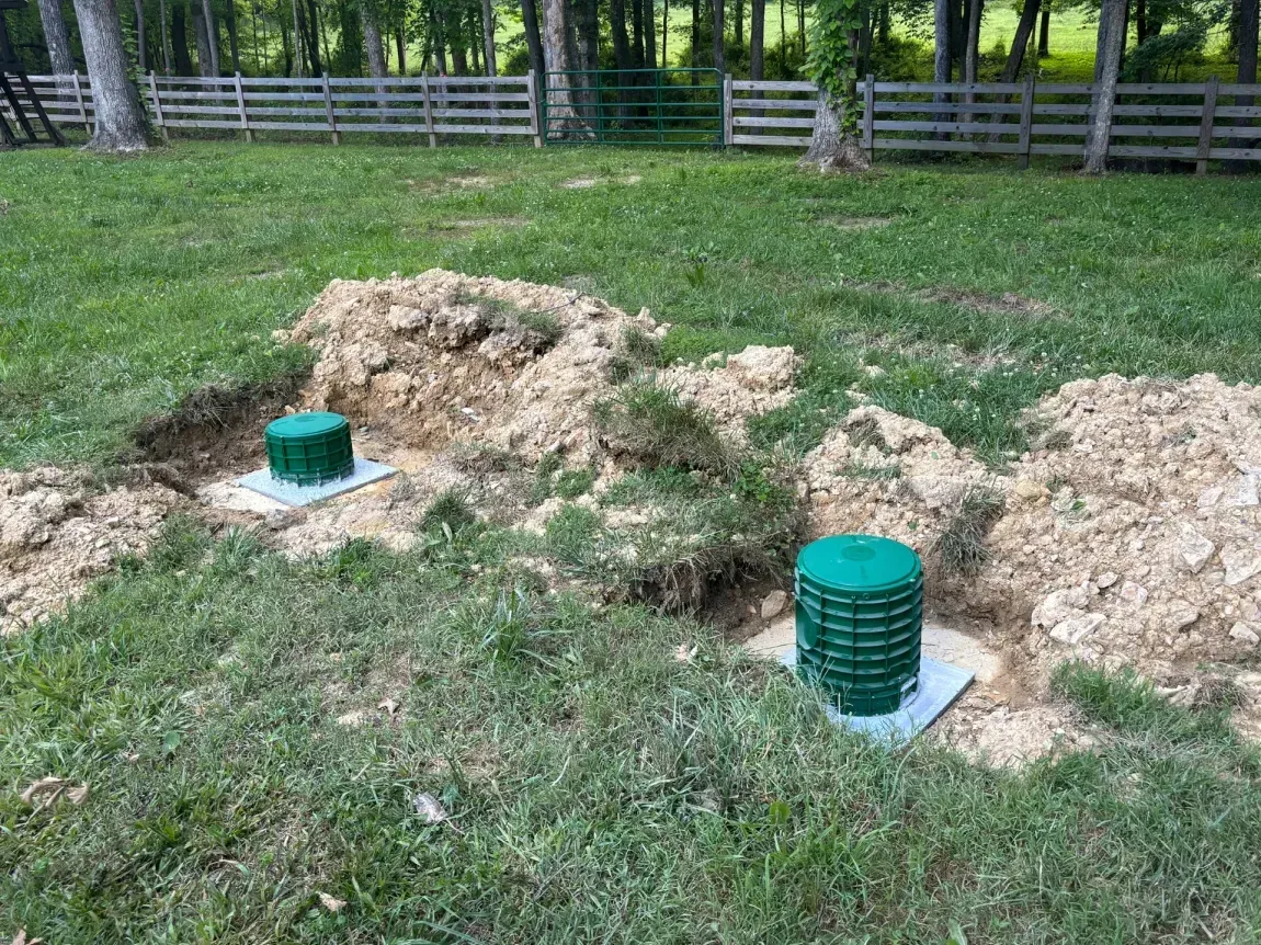 Two green septic tank lids in a grassy yard with dirt mounds and a wooden fence in back
