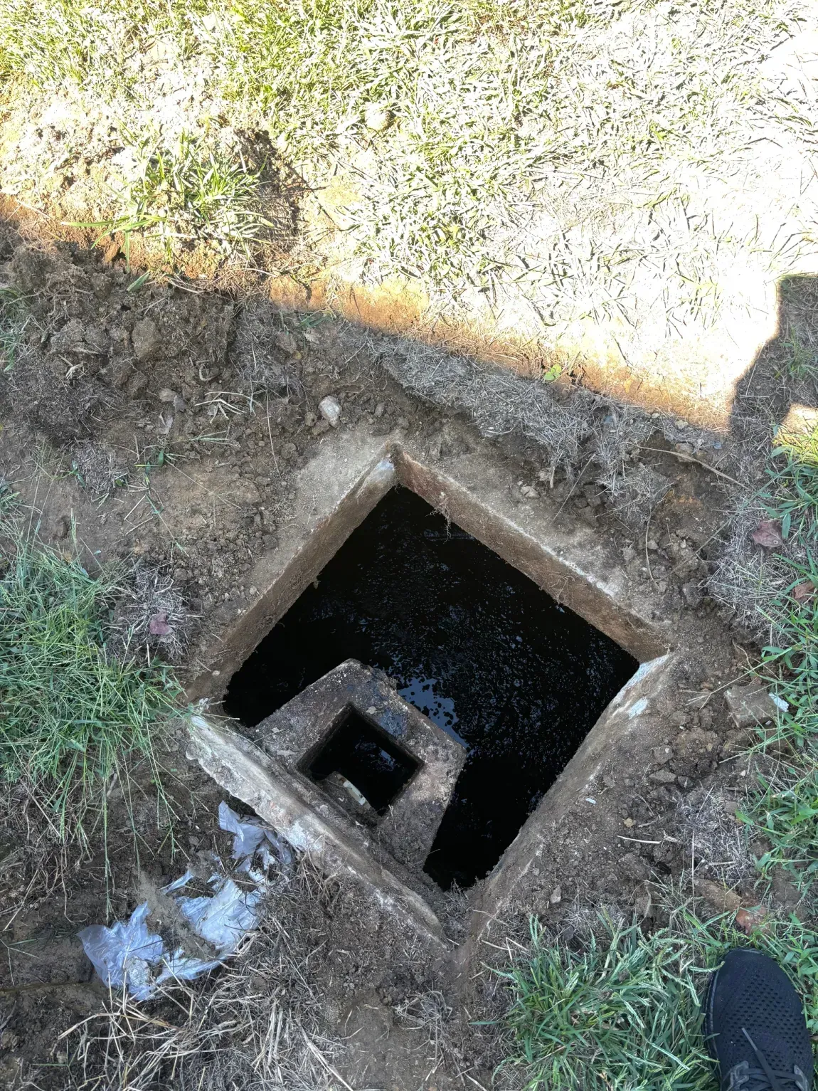 Open square utility hole in the ground with a smaller opening visible inside, near grass and dirt.