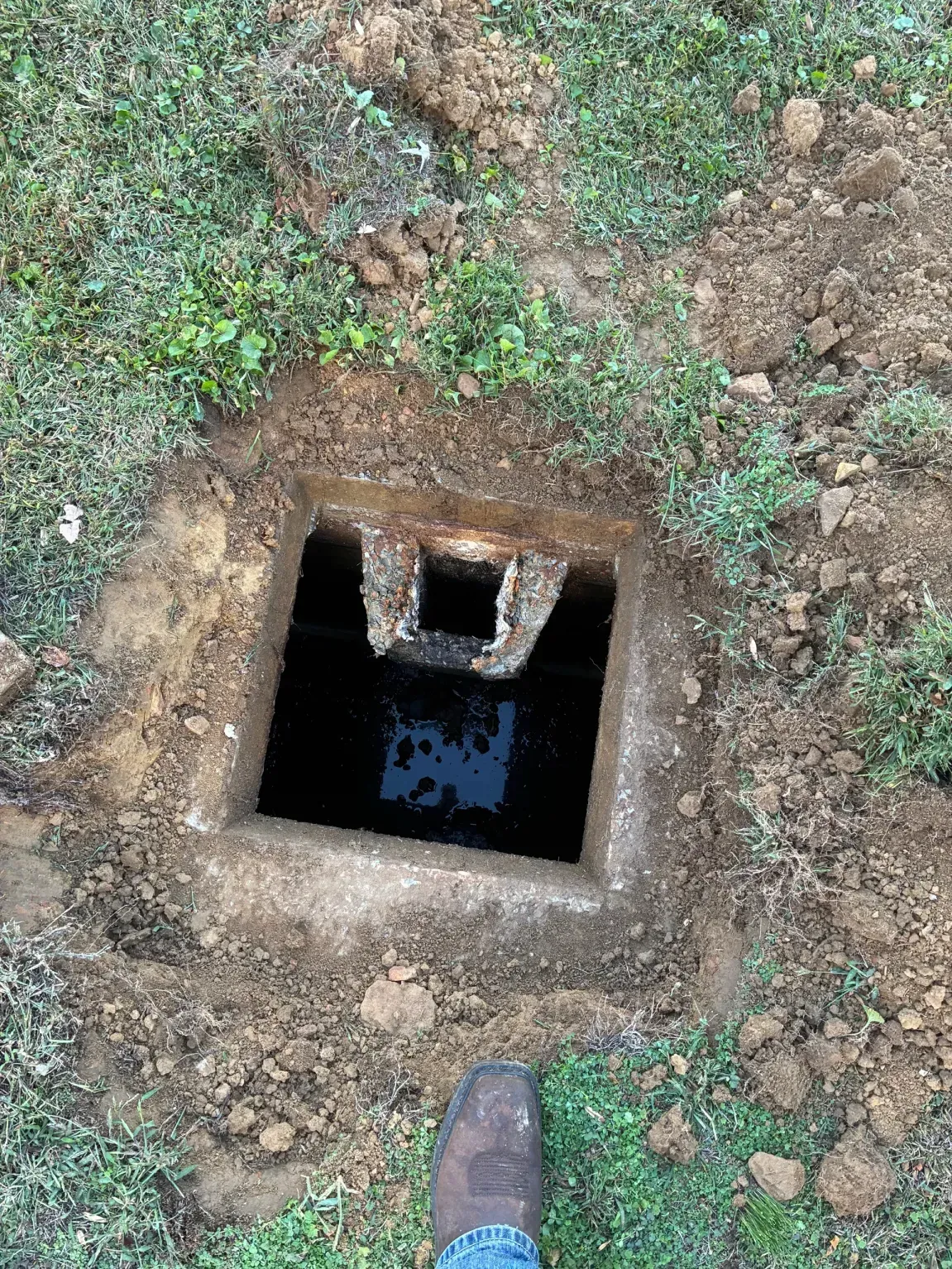 Open square pit in the ground with a dark opening and a pipe, surrounded by dirt and grass.