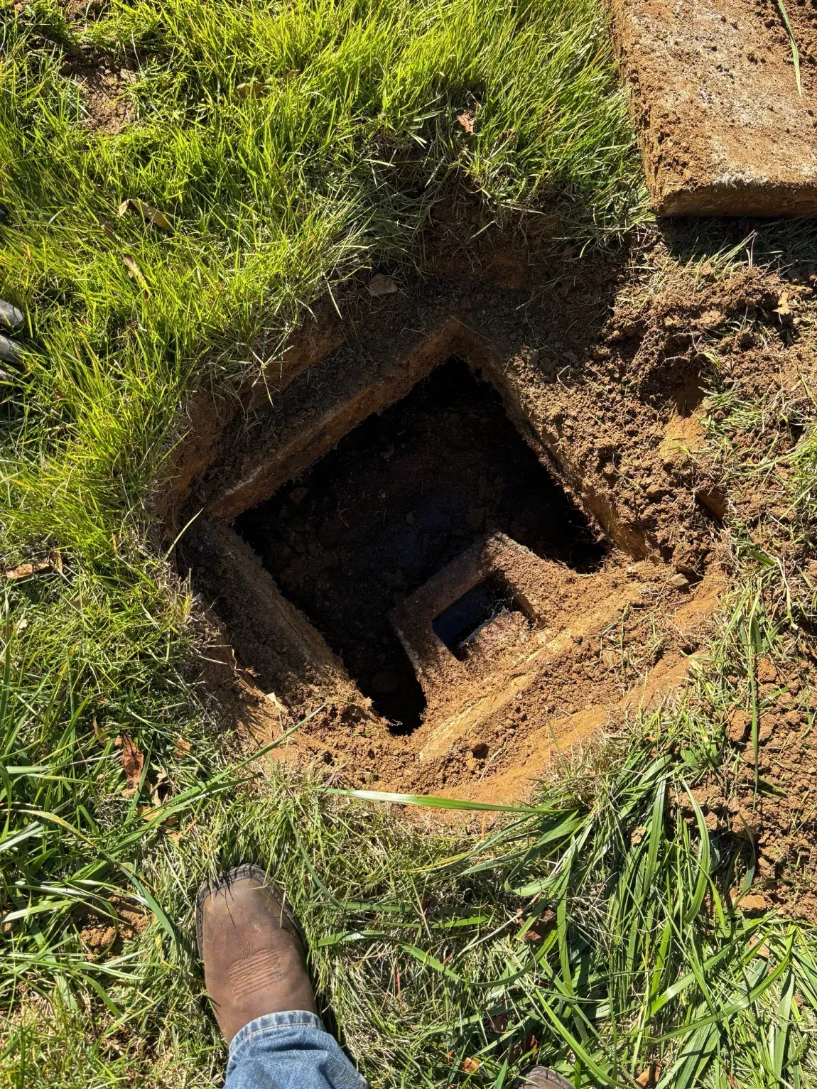Open pit in grassy ground with a dark square hole and a shoe at the bottom edge