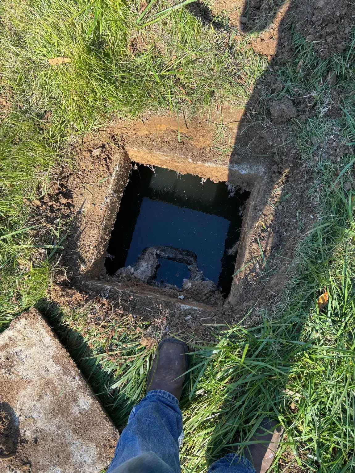 Open ground pit with dark water, seen from above beside a person’s leg and shoe.