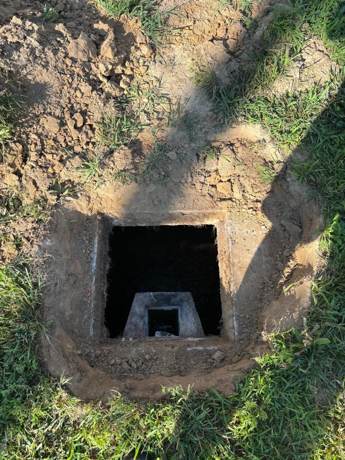 Open square pit in the ground with a smaller square structure inside, surrounded by dirt and grass.