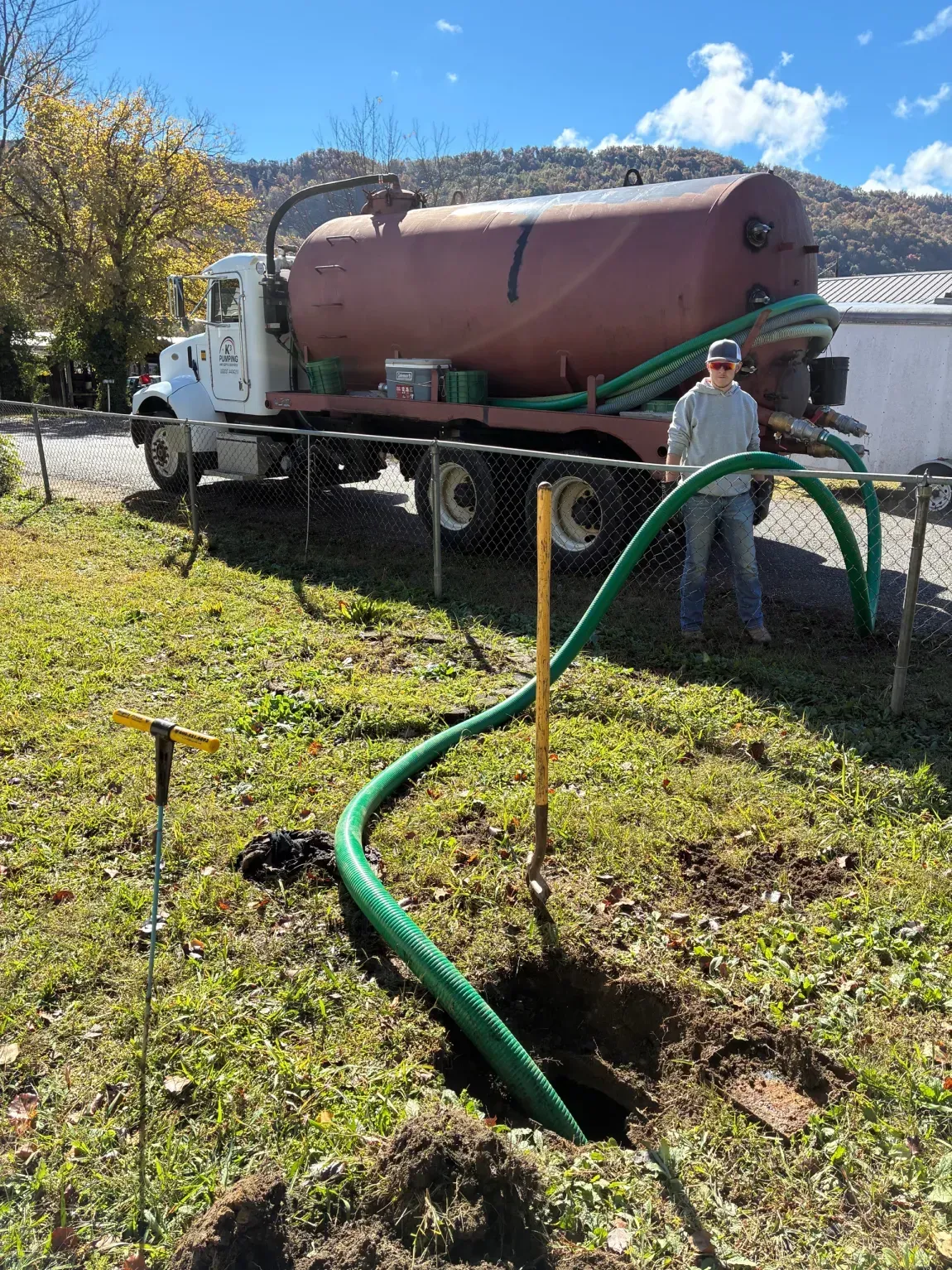 Worker by a white vacuum truck, pumping green hose in a sunny yard with a dirt hole in the foreground