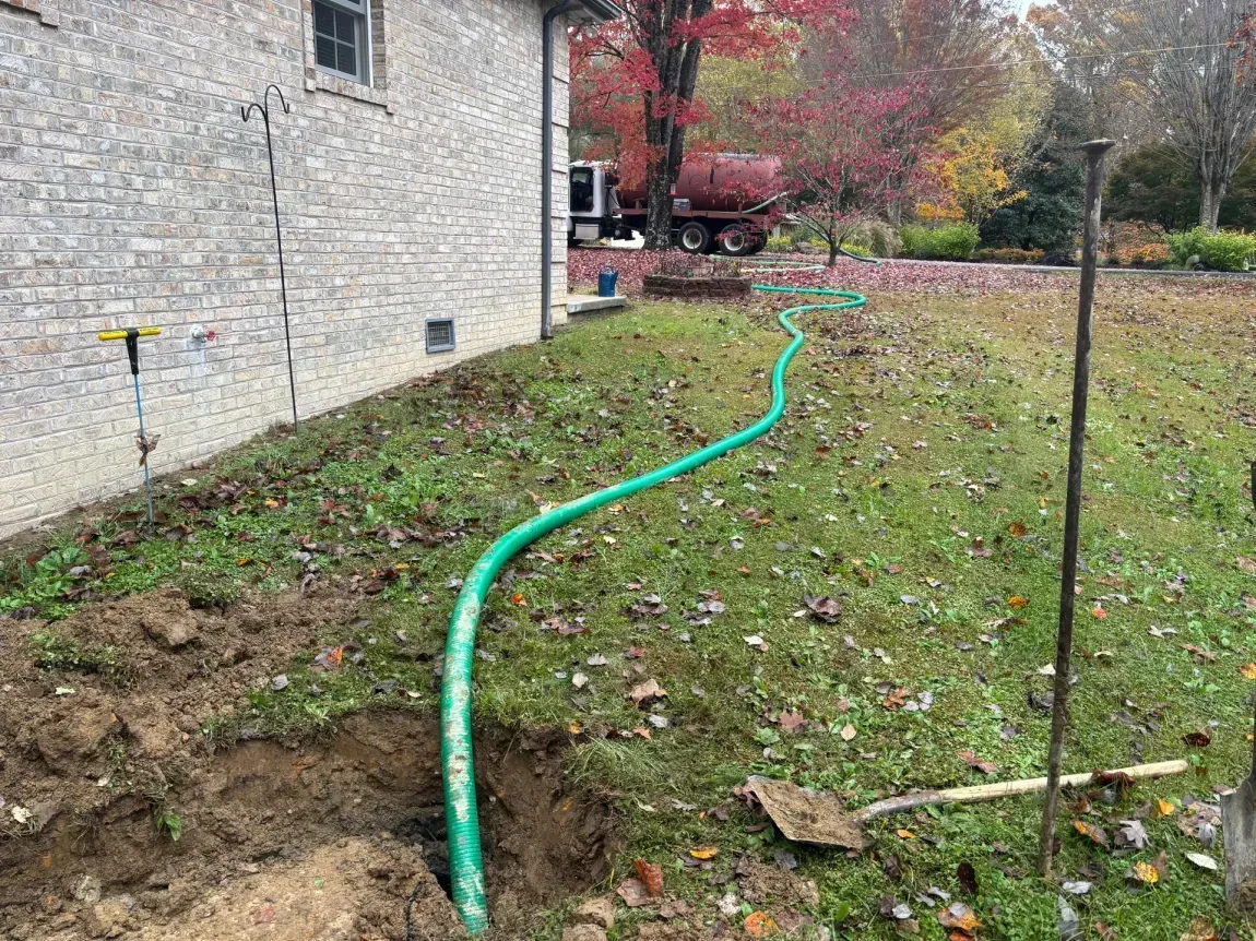 Green garden hose running along a muddy side yard beside a stone house in autumn.