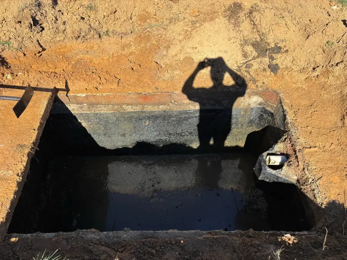 Shadow of a person standing beside a dark rectangular water basin in a sandy area.