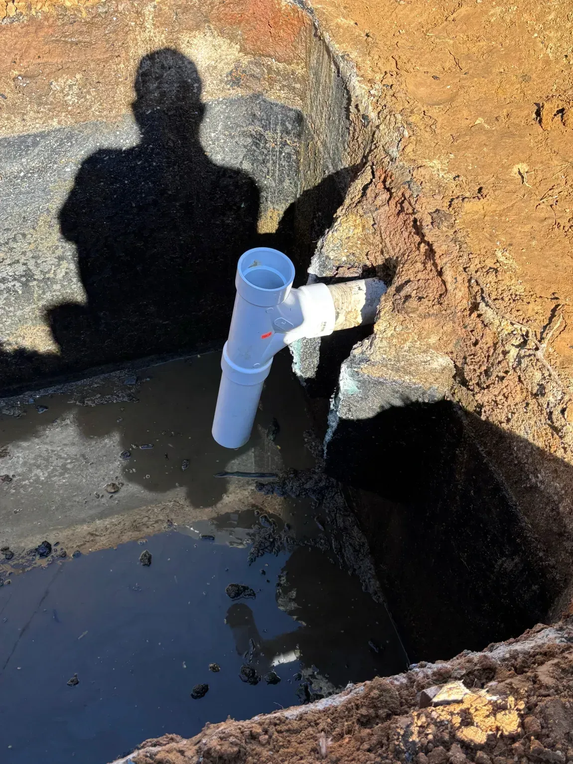 White PVC pipe in a muddy excavation trench beside a stone wall, with a person’s shadow on the wall.