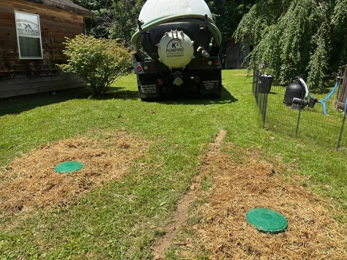 Two green utility covers in a grassy yard, with a large black tanker truck in the background.