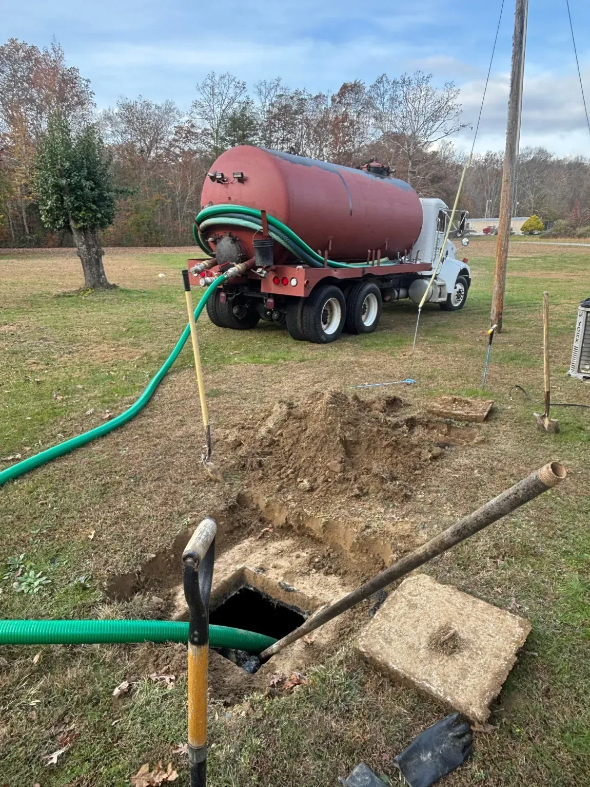Red tanker truck pumping into a green hose beside an open trench in a grassy yard