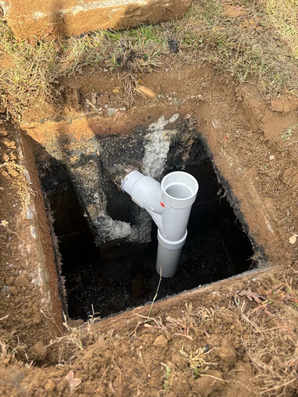 White PVC pipe fittings in a square dug hole in brown soil, with a black interior and rock edges.