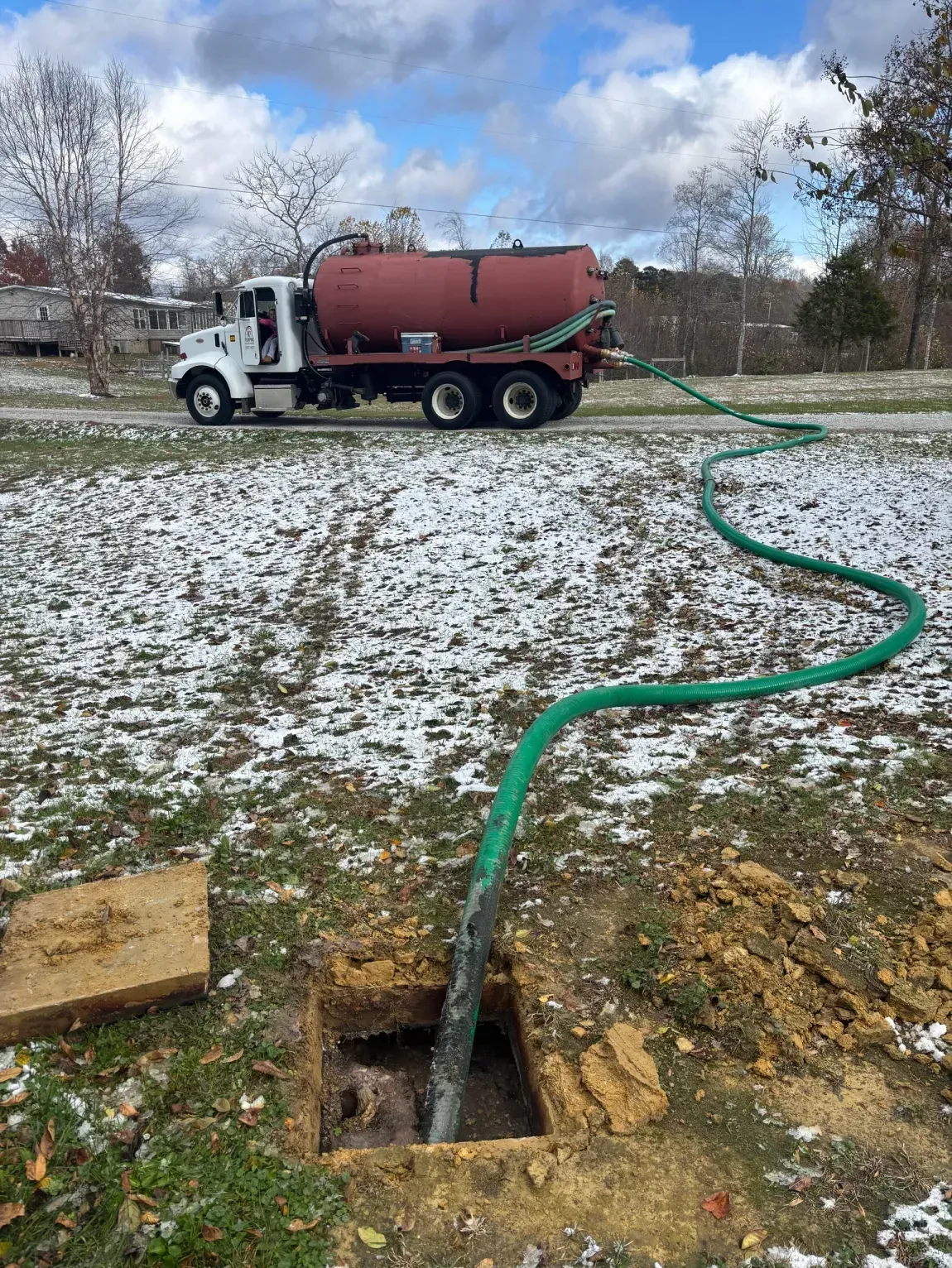 Tank truck pumping through green hose into an open ground pit in a snowy field