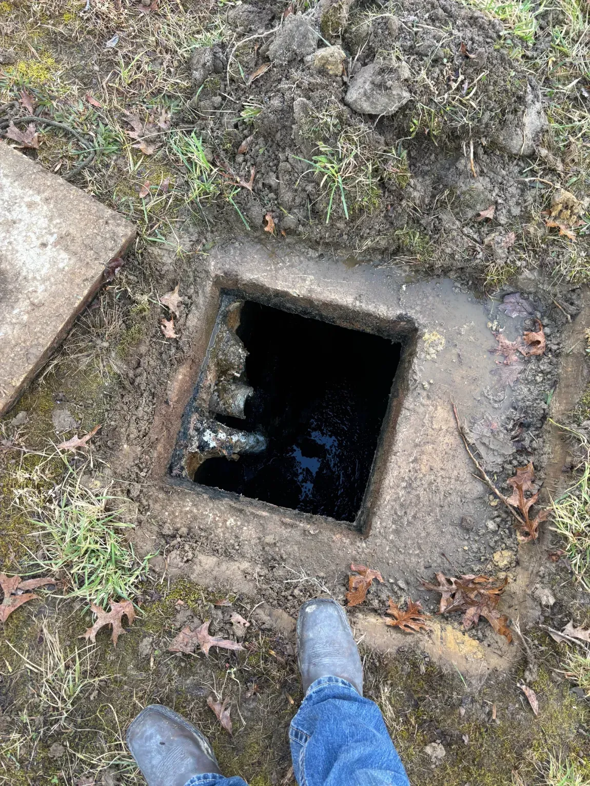 Open square utility hole in the ground, with a person's blue-jeaned feet nearby.