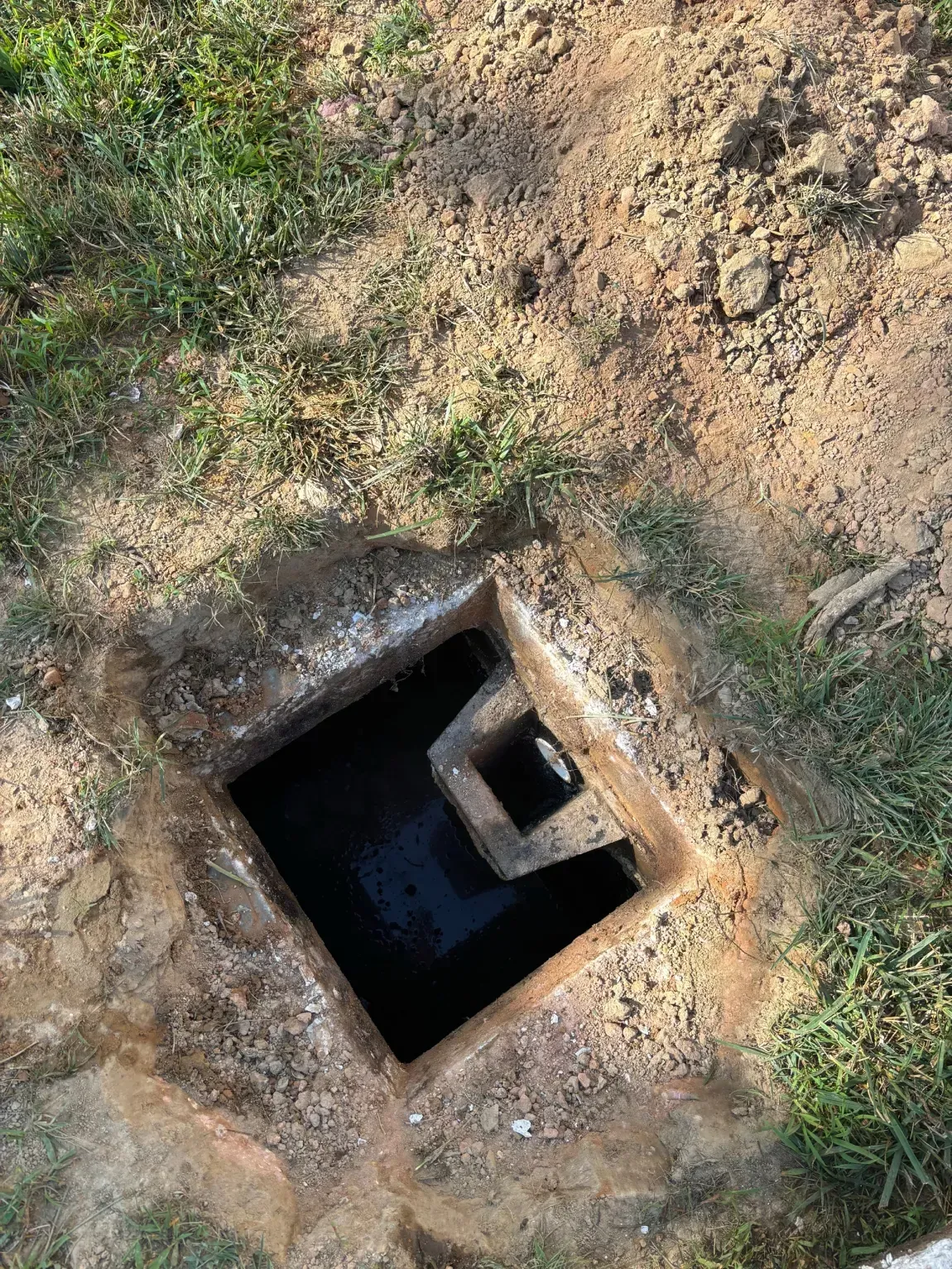 Open square concrete shaft in dirt and grass, with a dark hole and small inner ledge.