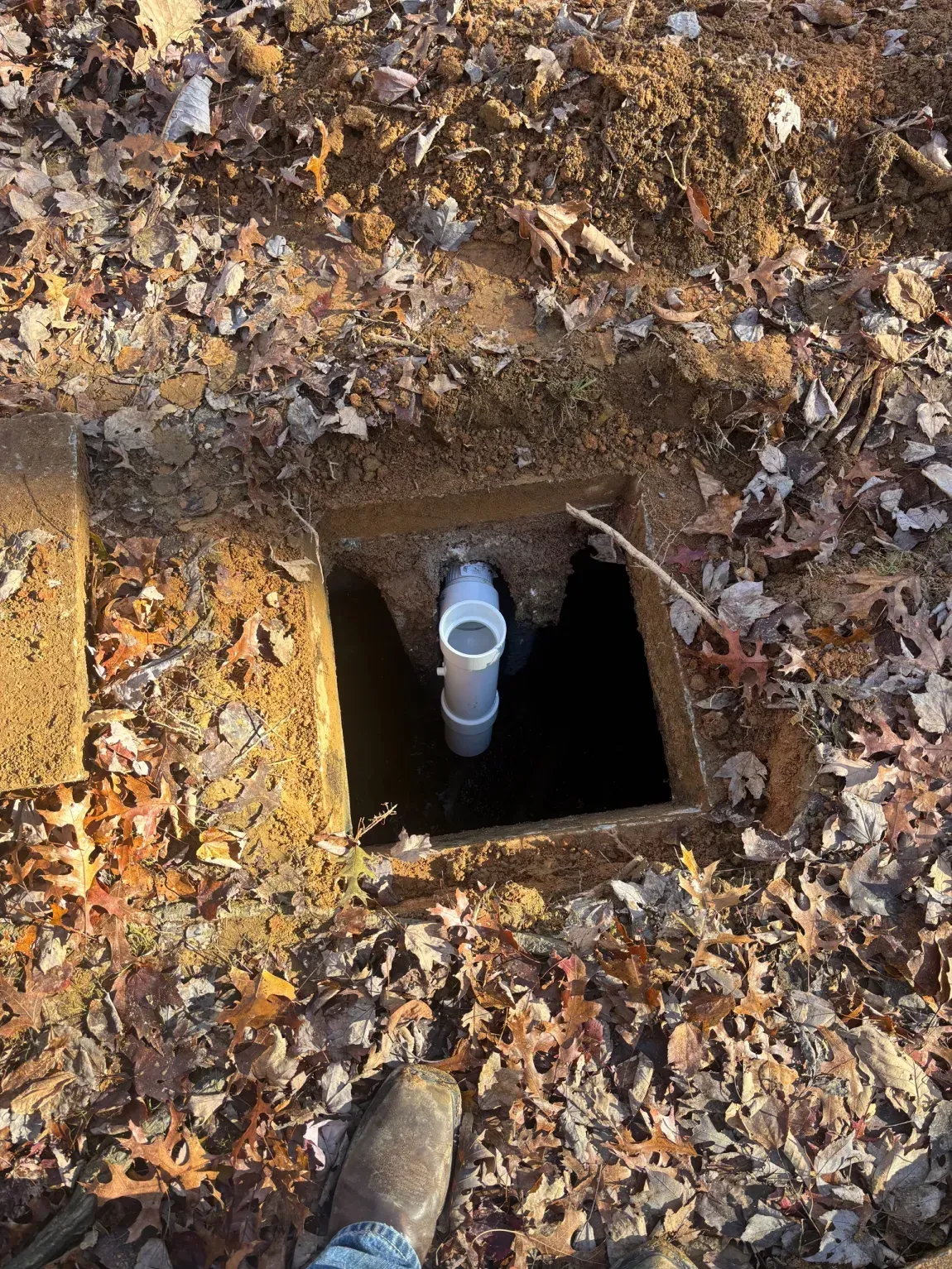 Open utility pit with a white pipe inside, surrounded by dirt and fallen leaves.