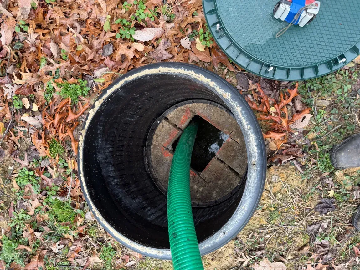 Open manhole with green hose feeding into a circular drain, surrounded by fallen leaves and a nearby cover