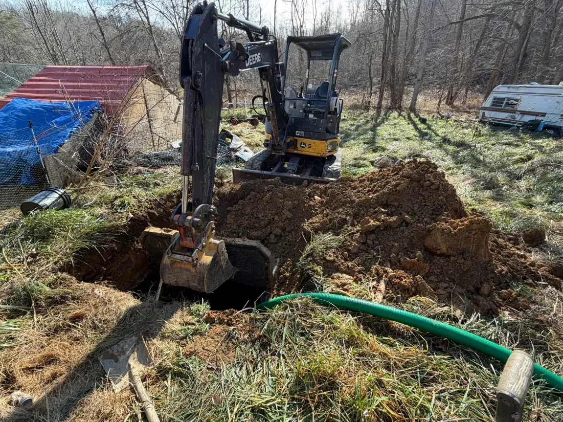 Excavator digging a trench in a grassy yard, with dirt piled nearby and a green hose in the foreground.