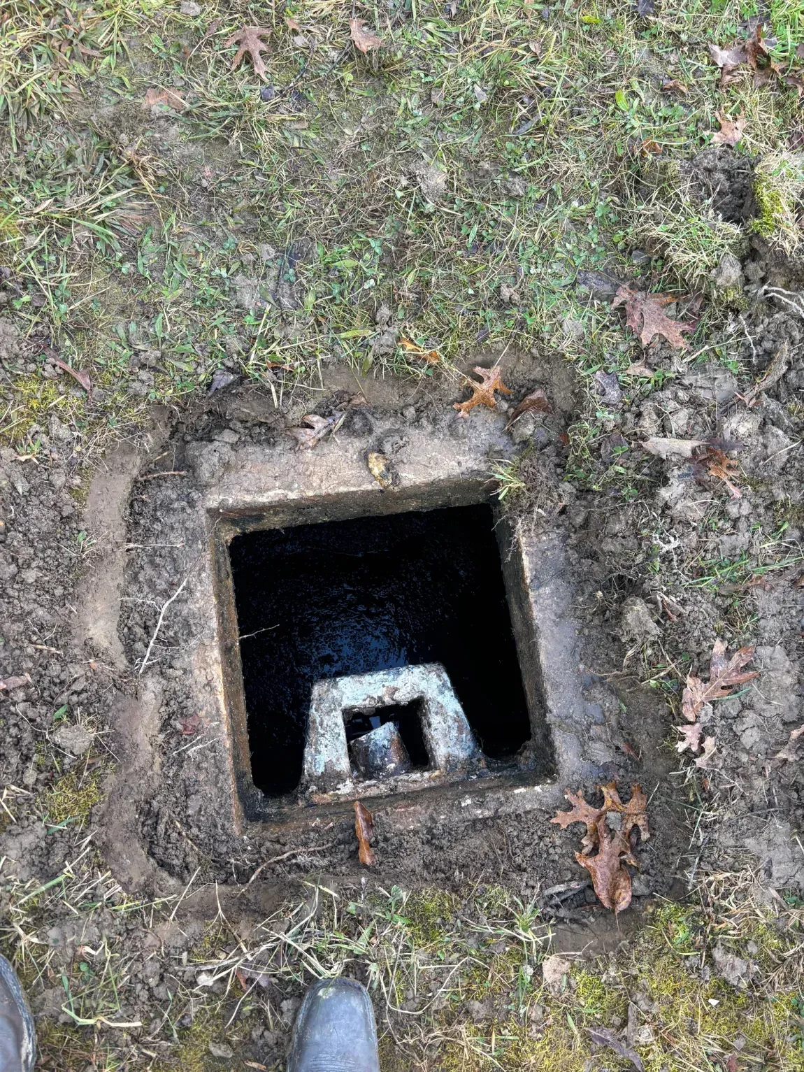 Open square utility hole in grassy ground with a metal bracket inside.