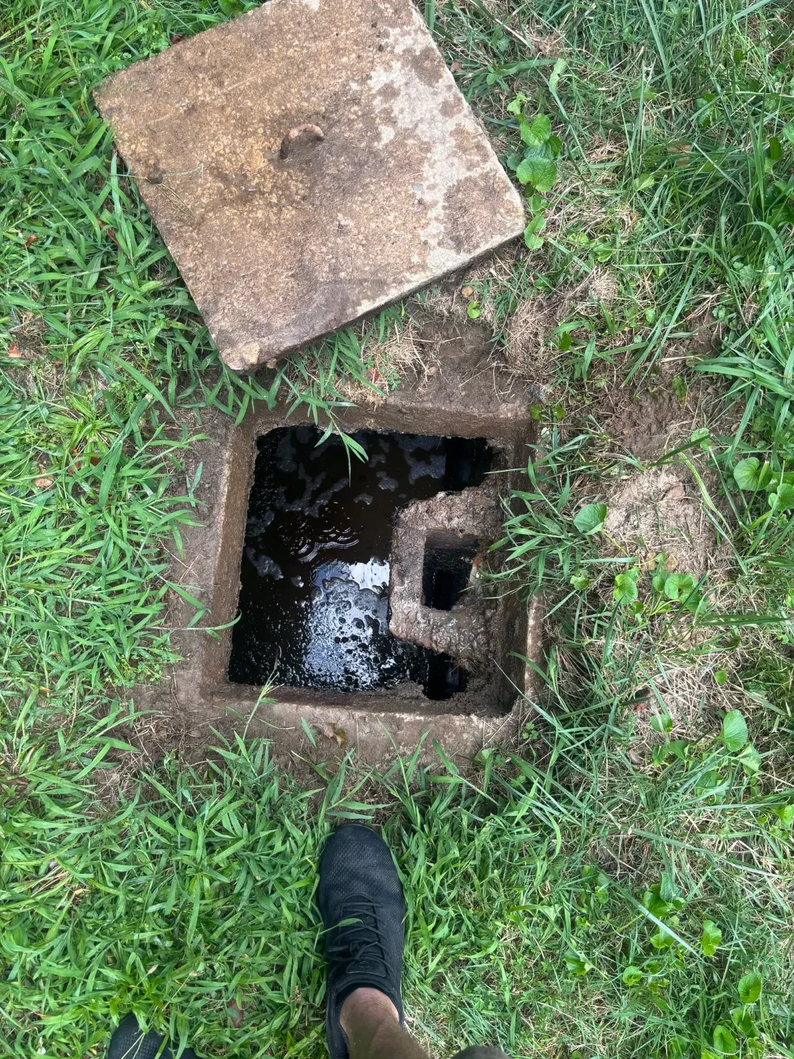 Open square hole in grass with concrete cover and a shoe at the edge