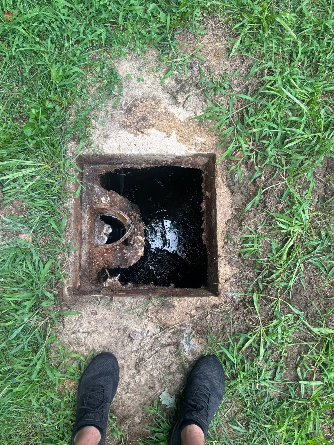Open square manhole in grass, with feet standing at the edge.
