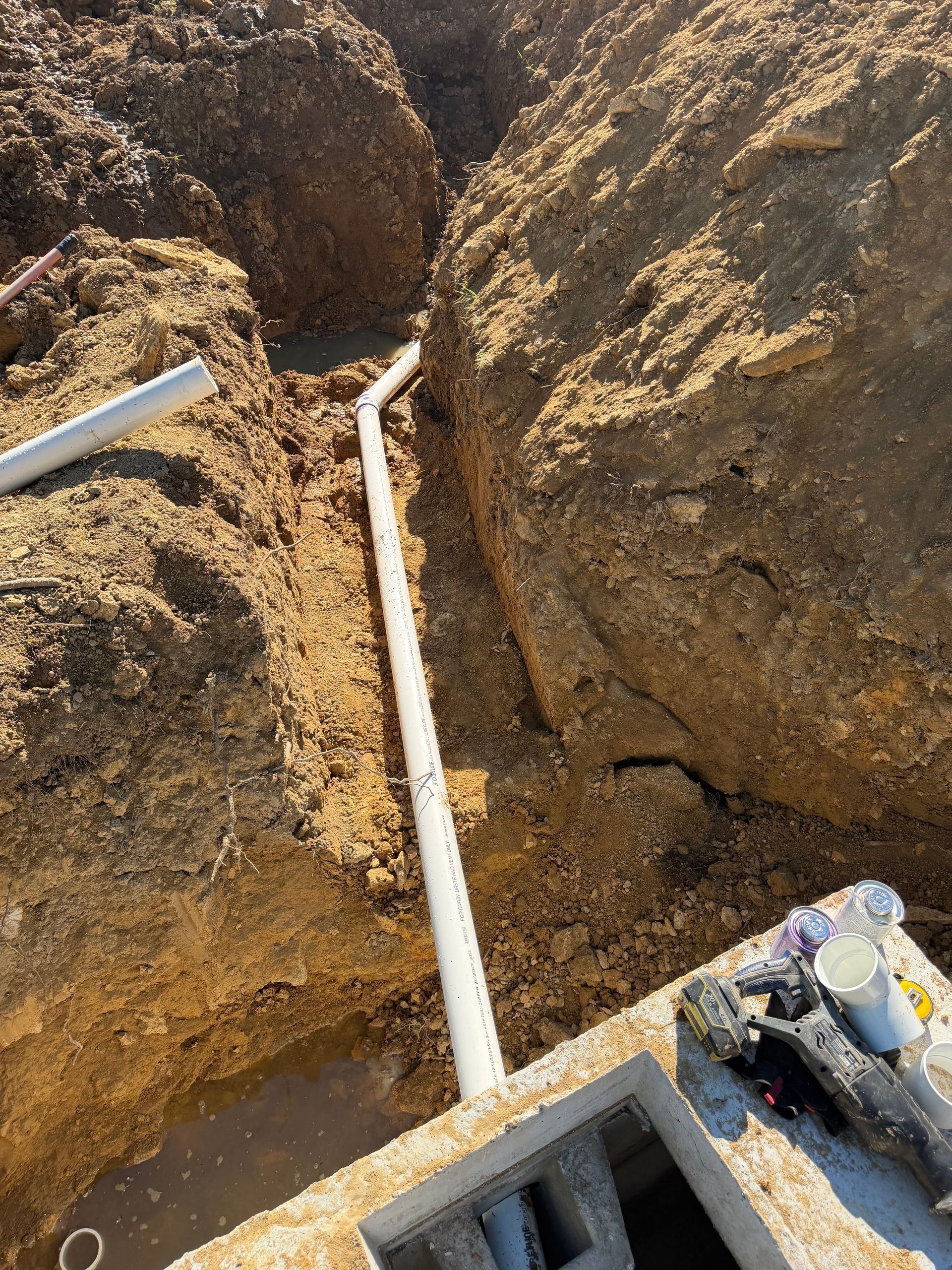 White PVC pipe running through a narrow dirt trench between rocky soil walls.