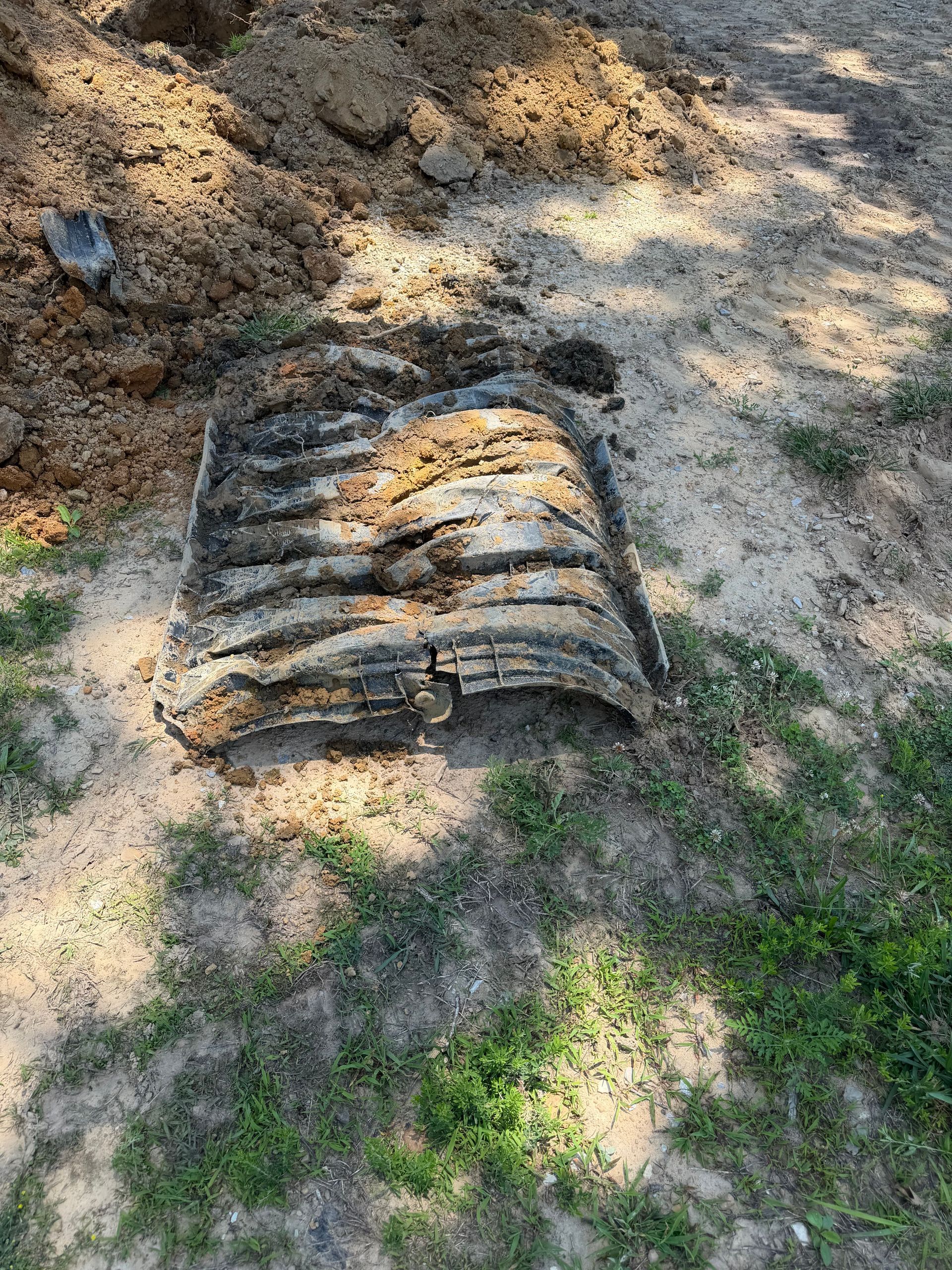 A rusty metal grating lies on a sandy dirt slope with scattered grass and sunlight.