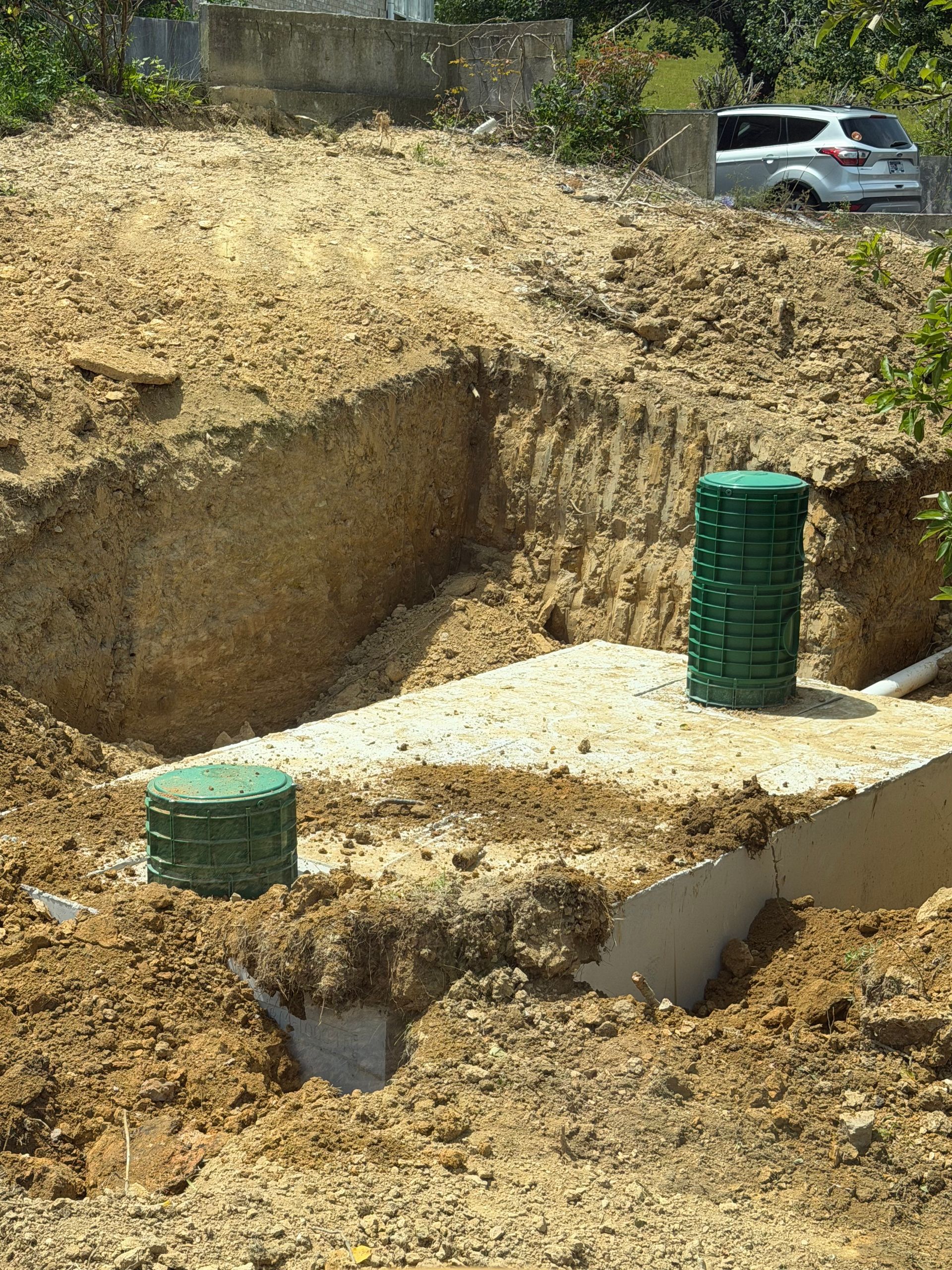 Construction pit with excavated dirt, concrete slab, and two green barrels near a parked SUV.