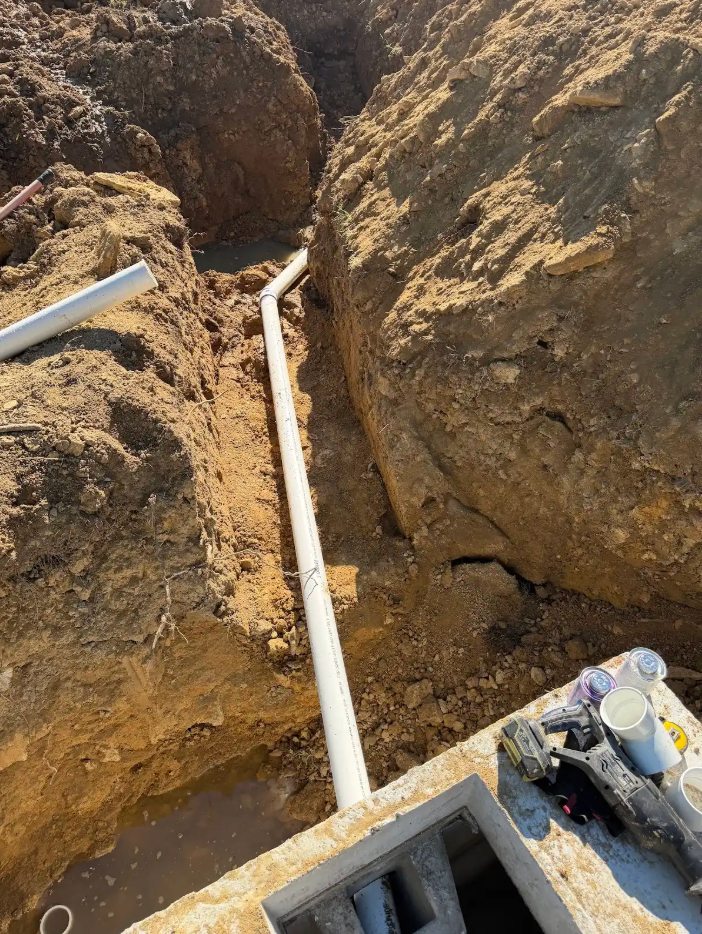 Trench with white PVC pipes laid between dirt walls beside construction equipment.