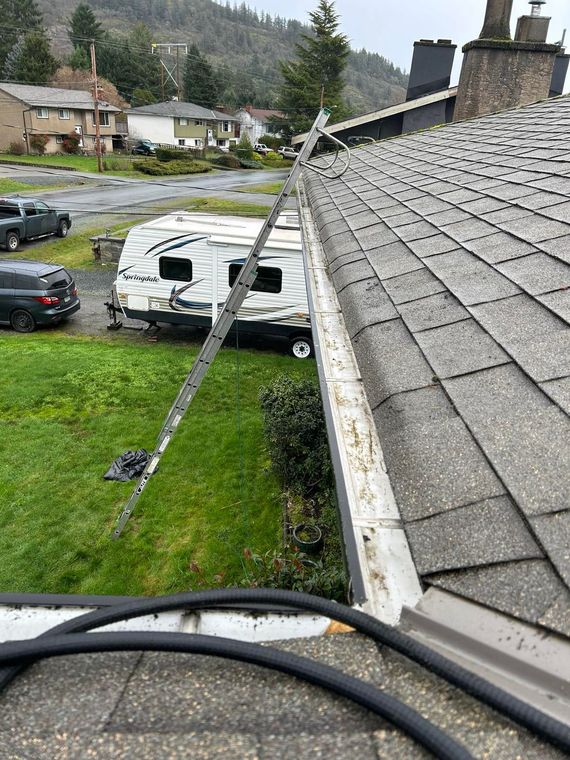 Ladder leaning against roof, cleaning gutters. A camper and vehicles are parked on a green lawn.