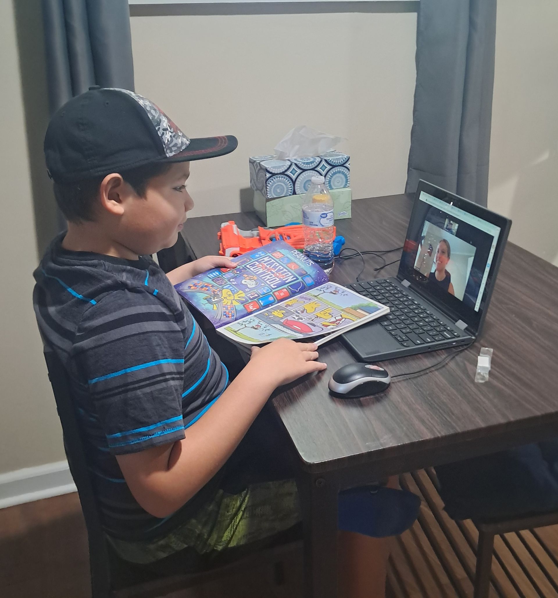 small boy at a laptop reading a comic book out loud to his reading tutor who appears on the computer screen