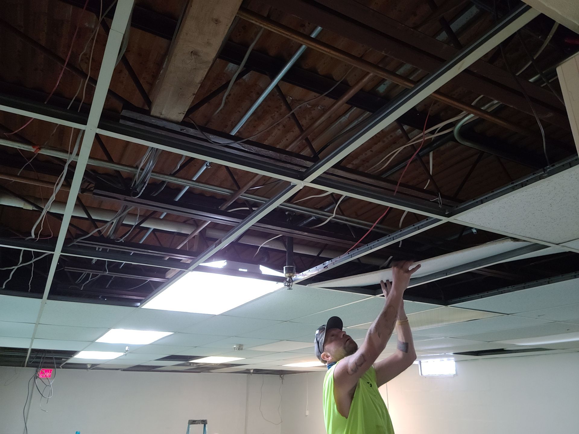 A person in a neon green shirt working on a partially demolished ceiling. Exposed beams and wires visible.