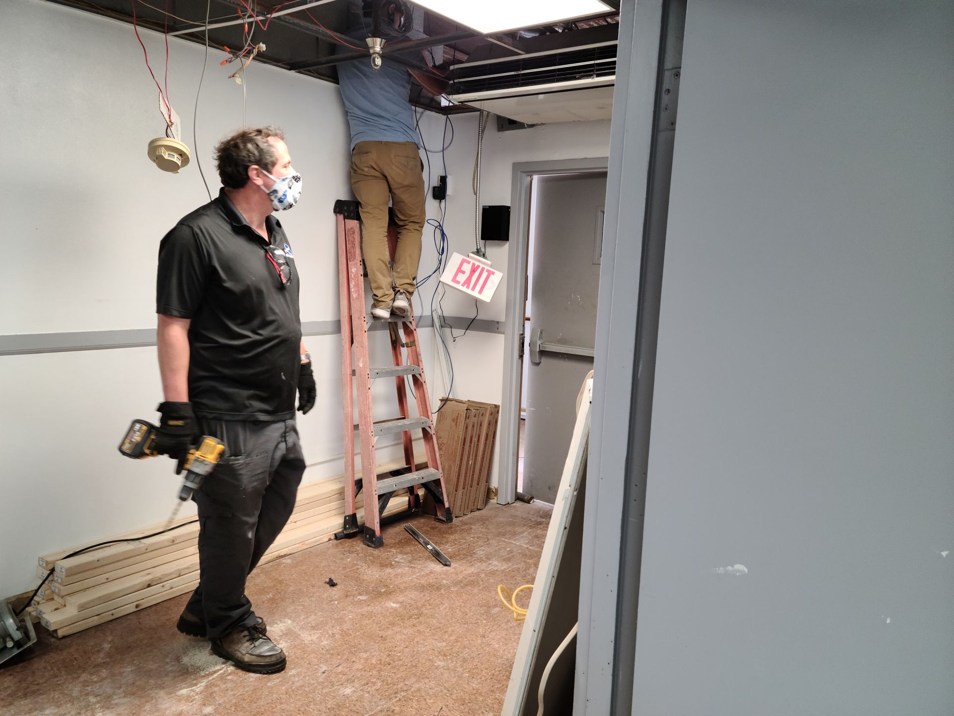 Two men renovating a room: One on a ladder working on wires, the other holding a power tool.