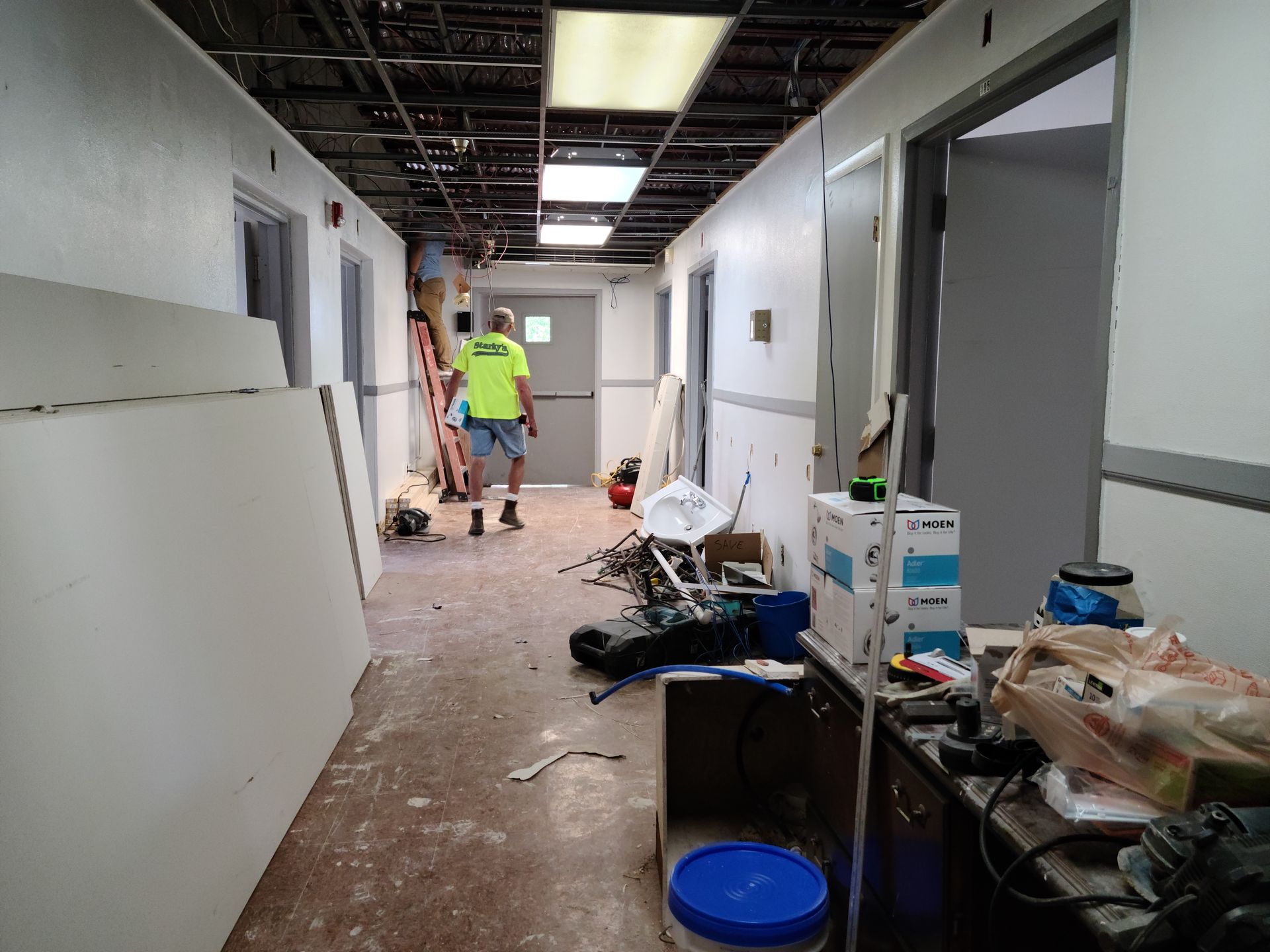 Hallway under construction with a worker on a ladder, drywall and supplies scattered.