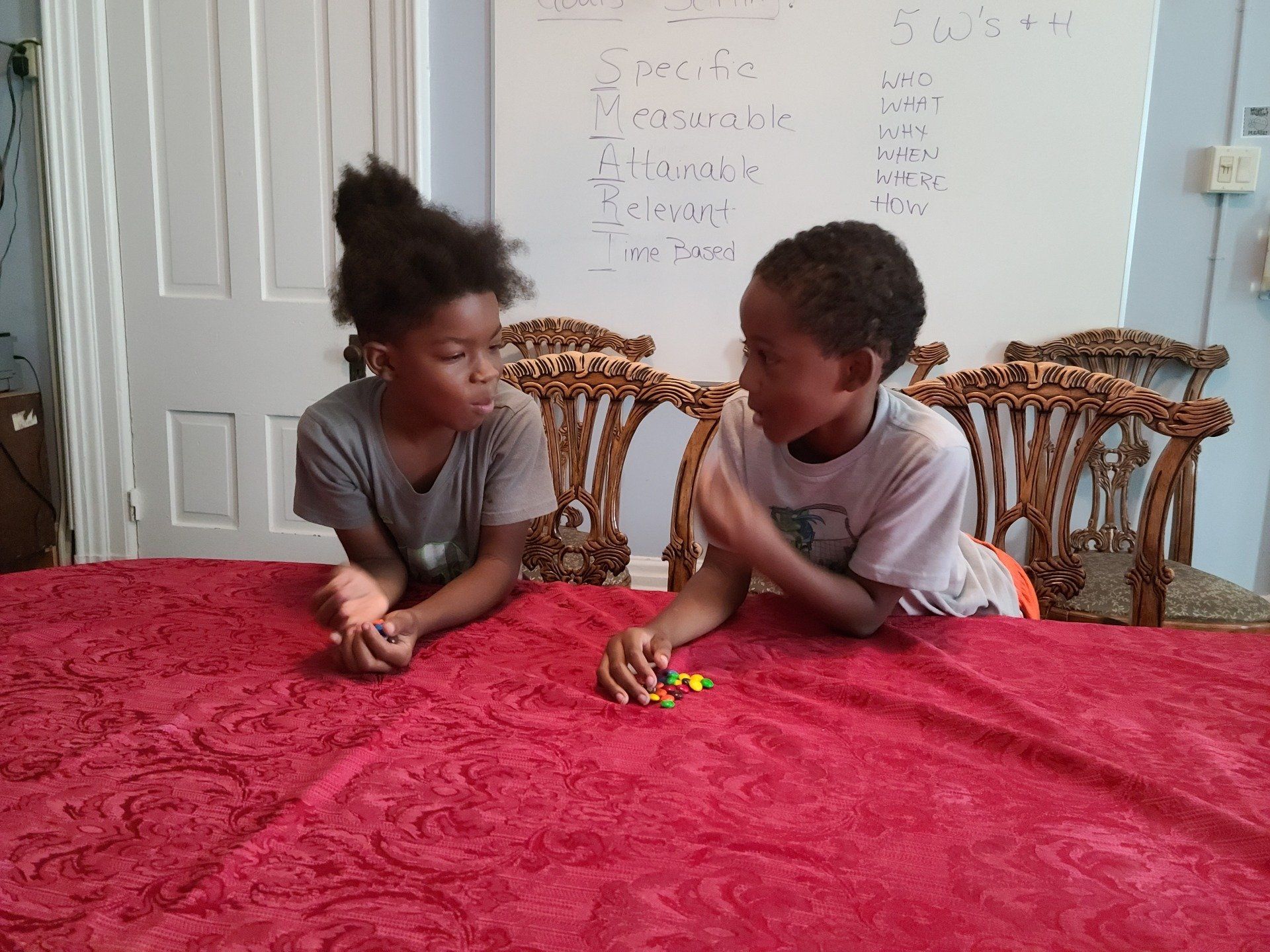Two children looking at each other over a red tablecloth, with a whiteboard in the background.