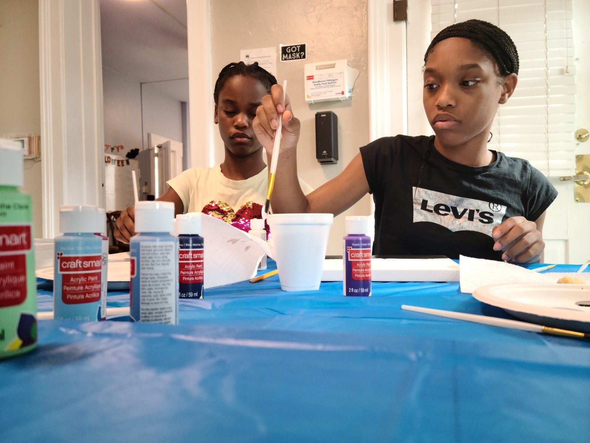 Two young Black girls painting at a table covered in blue. Paint and brushes are visible.