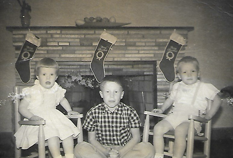Three children pose by a fireplace, Christmas stockings hung above. Two girls sit, one boy sits crossed-legged in front of the fire.